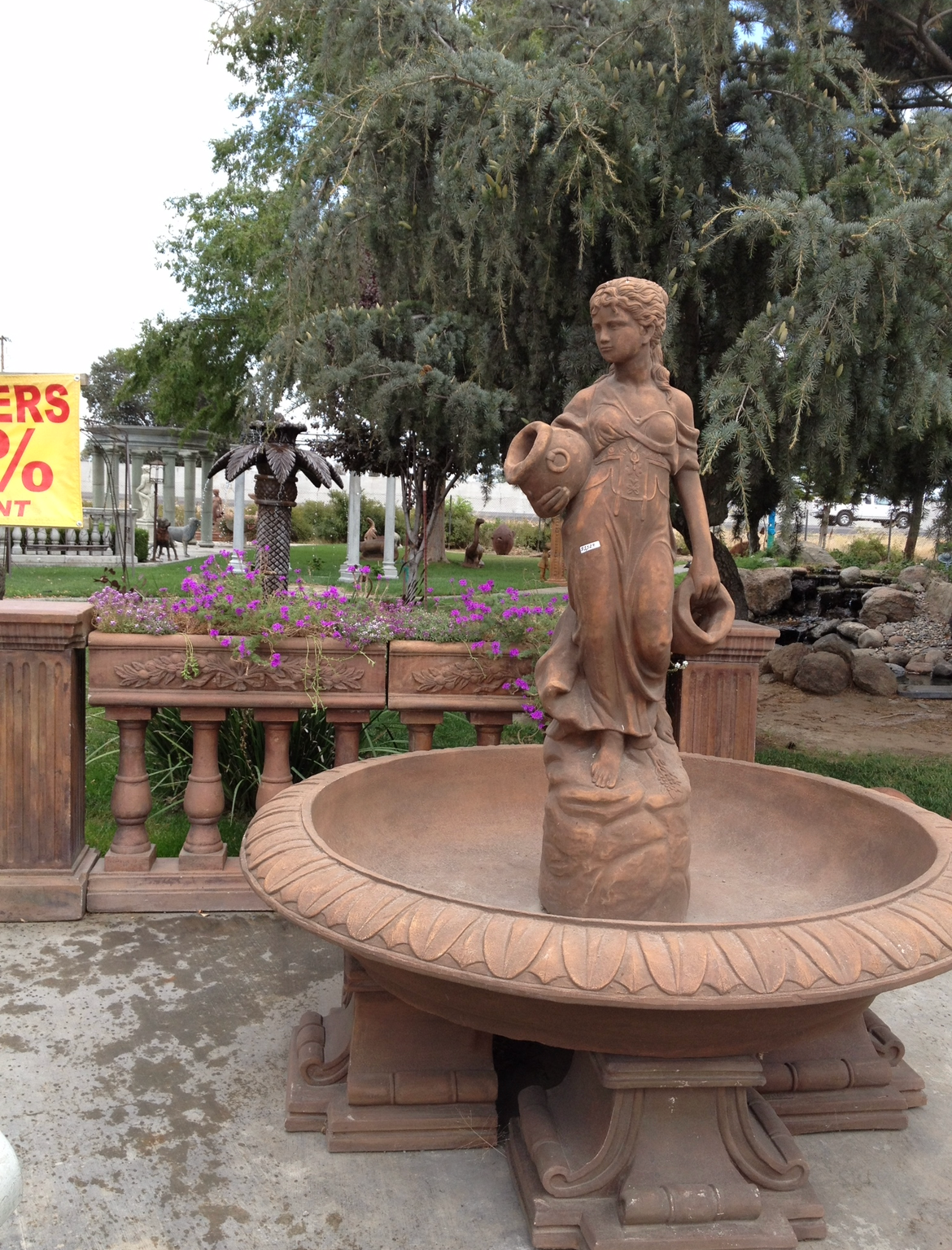 Stone fountain statue in a garden with flowers, trees, and a metal fence in the background