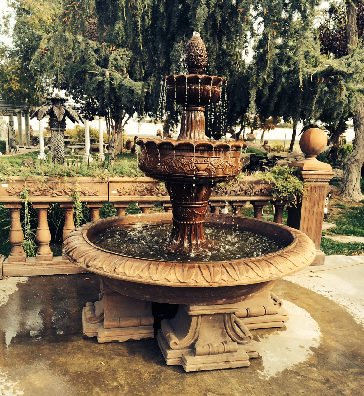 Ornate stone fountain in a garden, with trees and a low wooden fence in the background.