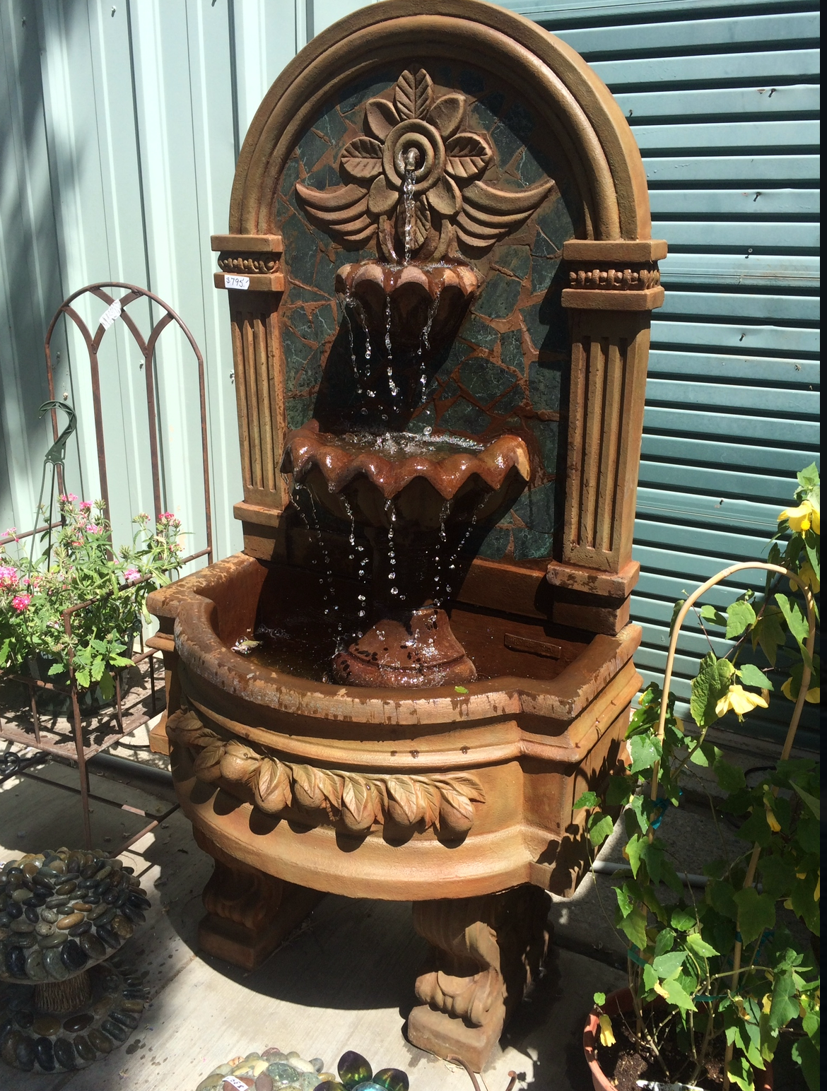 Ornate brown garden fountain with tiered basins beside a green shuttered wall and plants