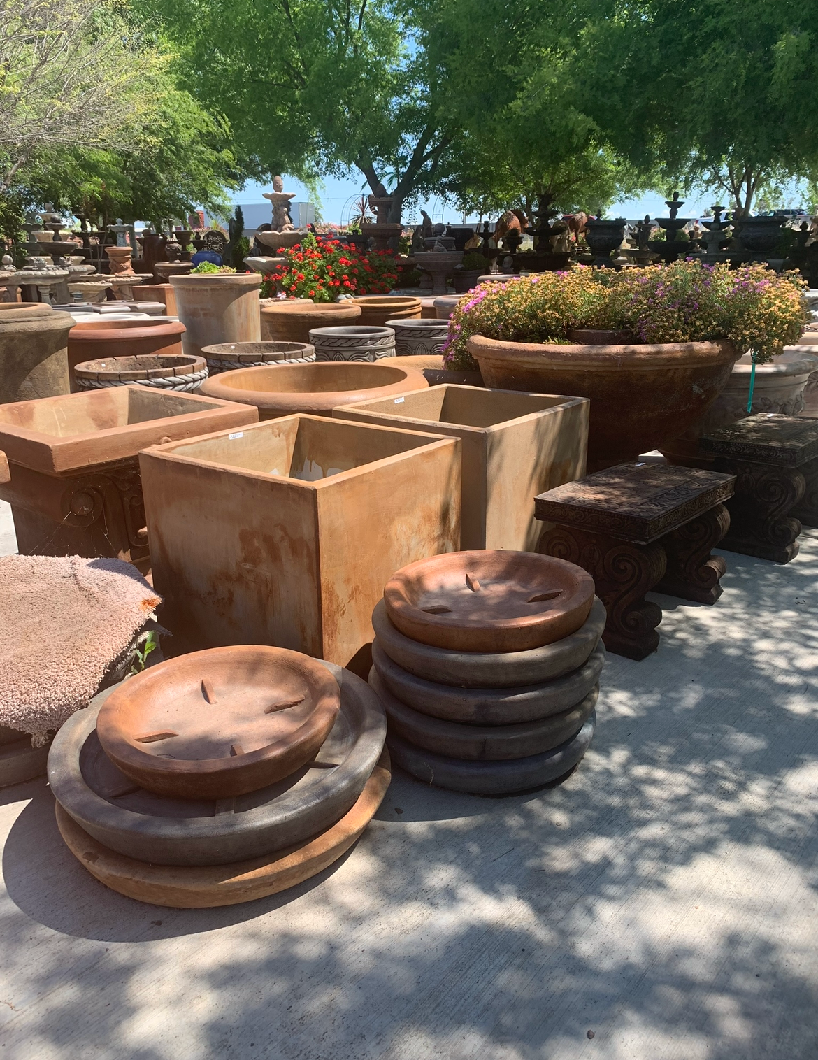 Outdoor garden display of rustic stone planters and stacked terracotta pots under trees in bright sunlight