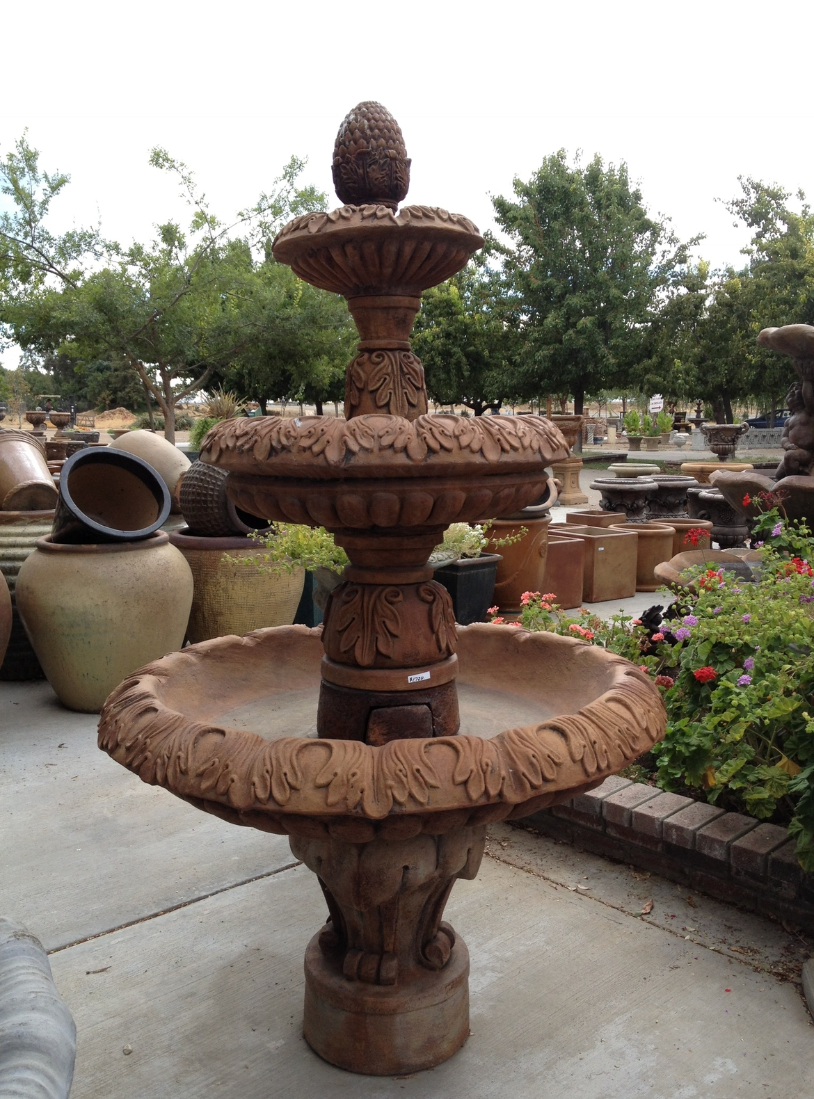 Ornate tiered stone fountain in a garden, with a pinecone finial and surrounding pottery.