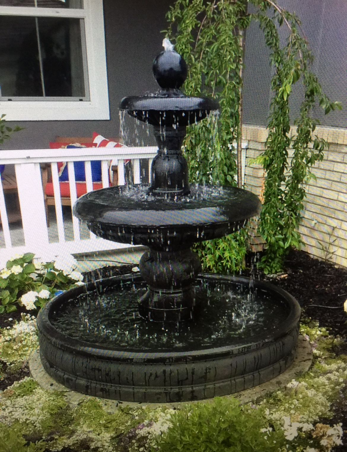 Black tiered garden fountain spraying water in a backyard beside a white porch.