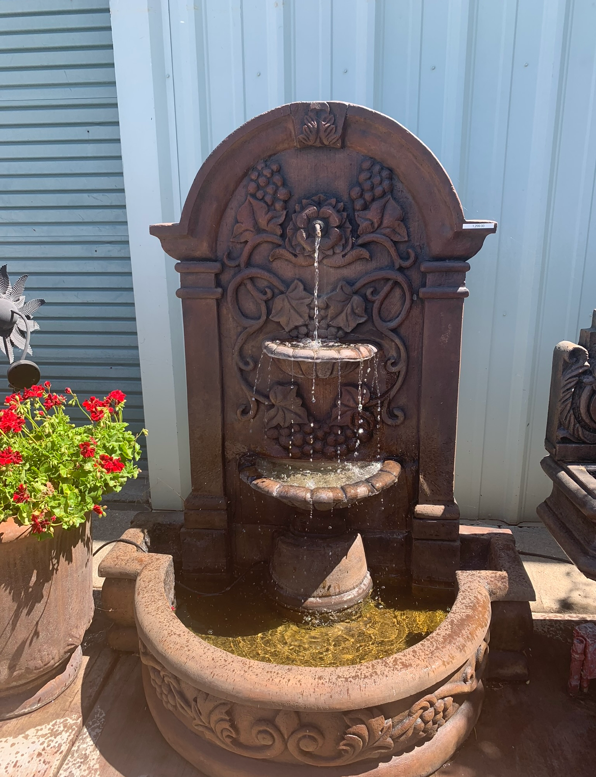 Ornate outdoor stone fountain with water flowing into a carved basin beside a flower pot and metal wall