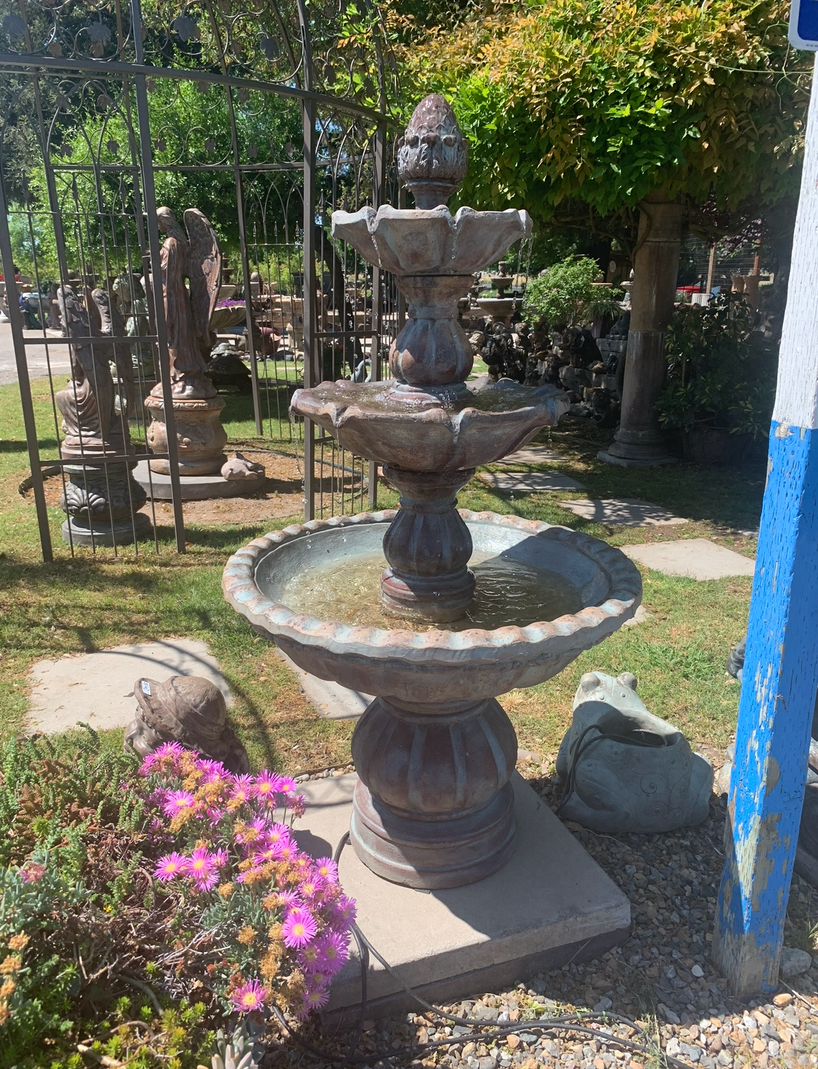 Three-tiered stone fountain in a garden, surrounded by flowers and greenery.