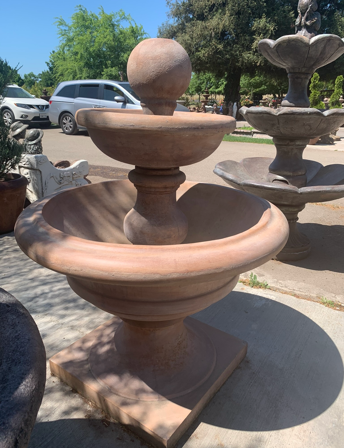 Large terracotta tiered fountain outdoors in a sunny garden, with parked cars and trees in the background
