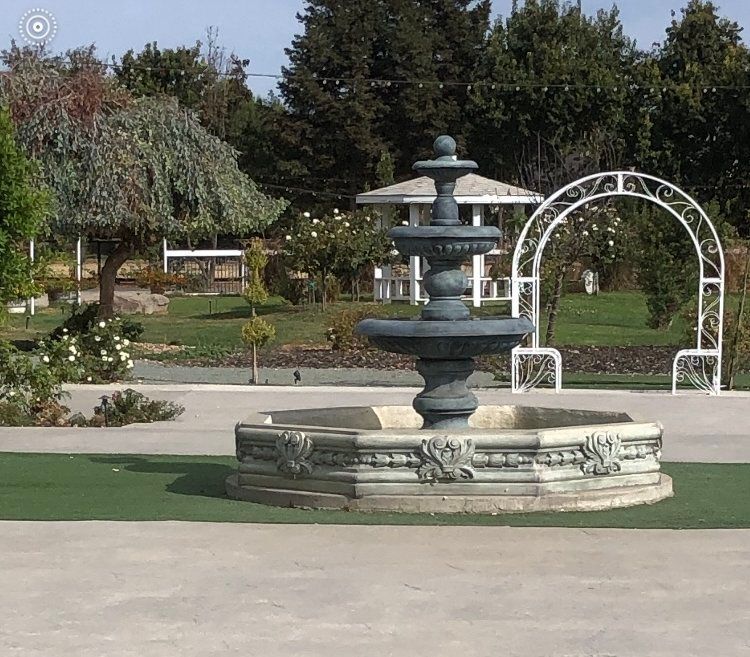 Decorative tiered fountain in a landscaped garden with trees and a white archway.
