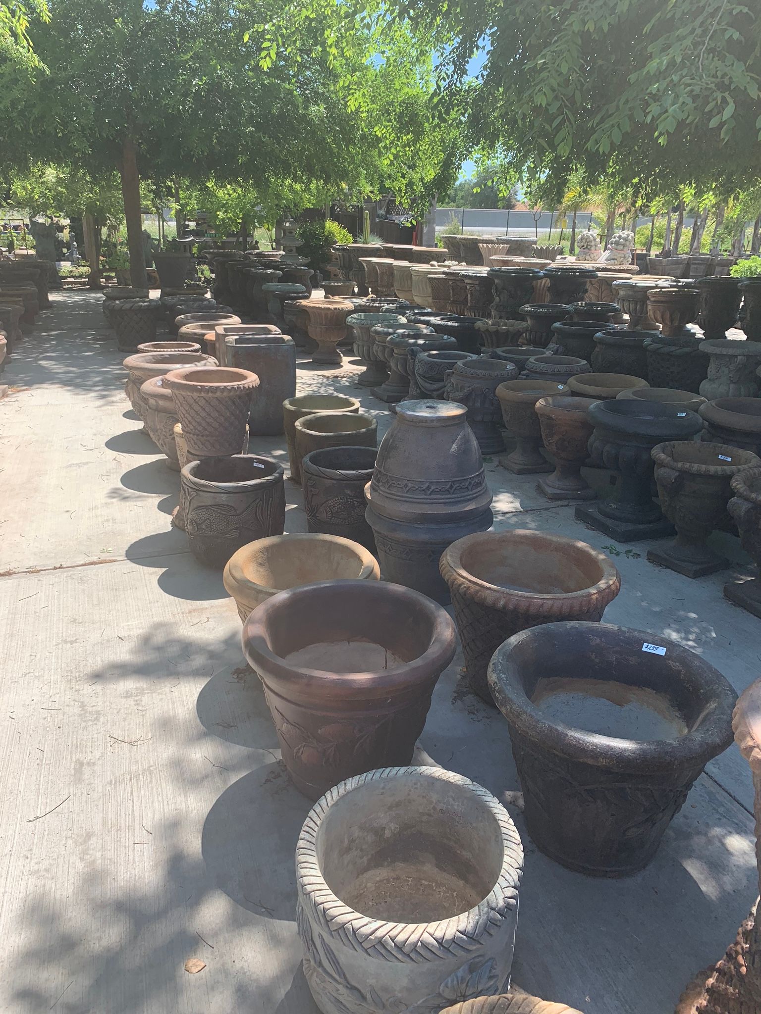Outdoor market display of large clay pots and planters on a paved path under leafy trees.