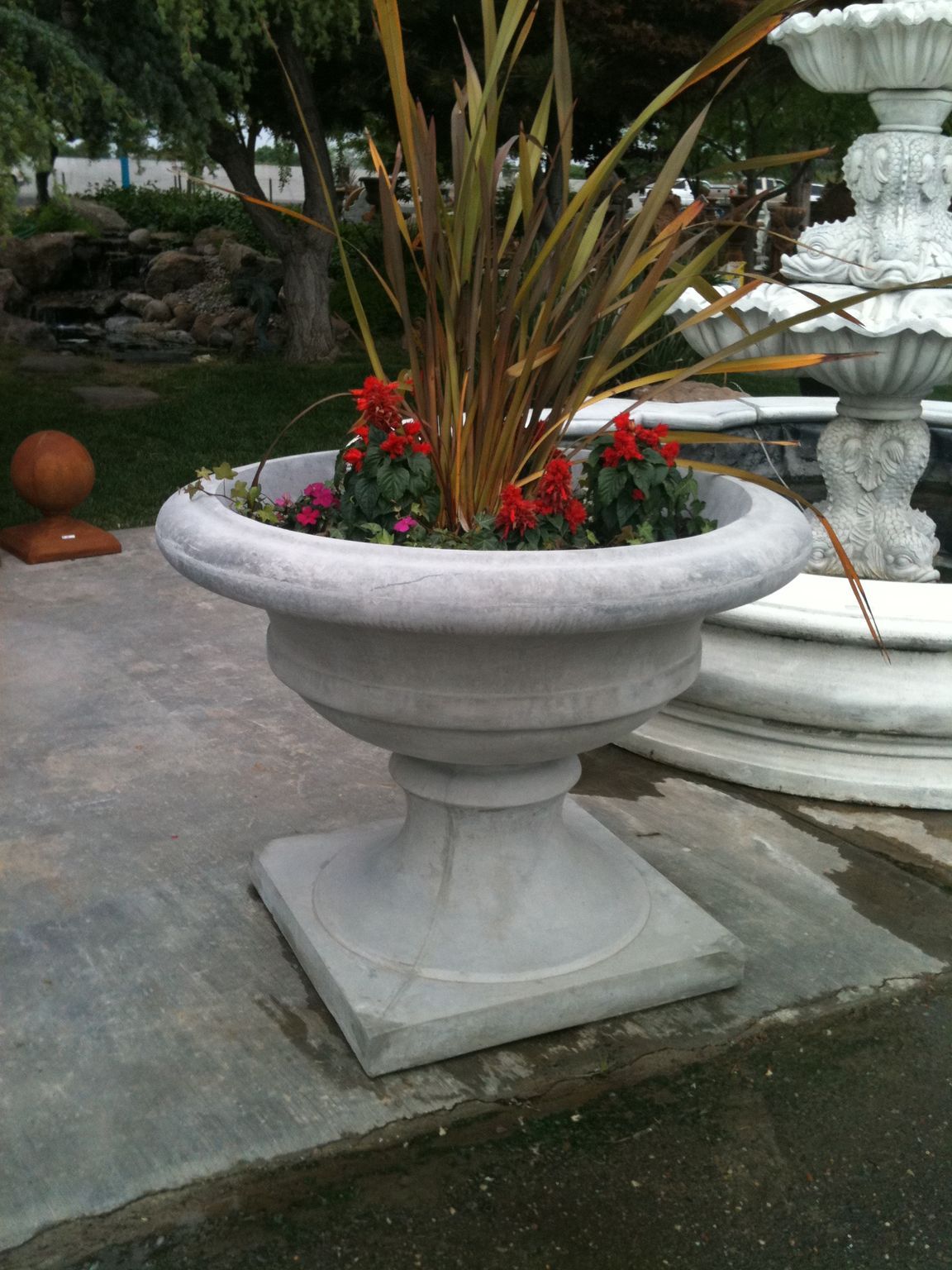 Large white stone planter with red flowers and tall grasses beside a fountain in a garden