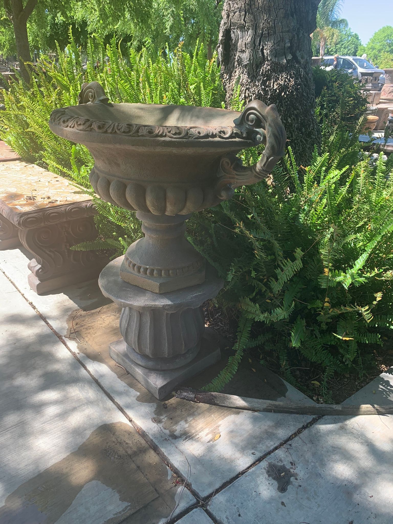 Stone urn planter beside a sidewalk, surrounded by green shrubs and dappled sunlight