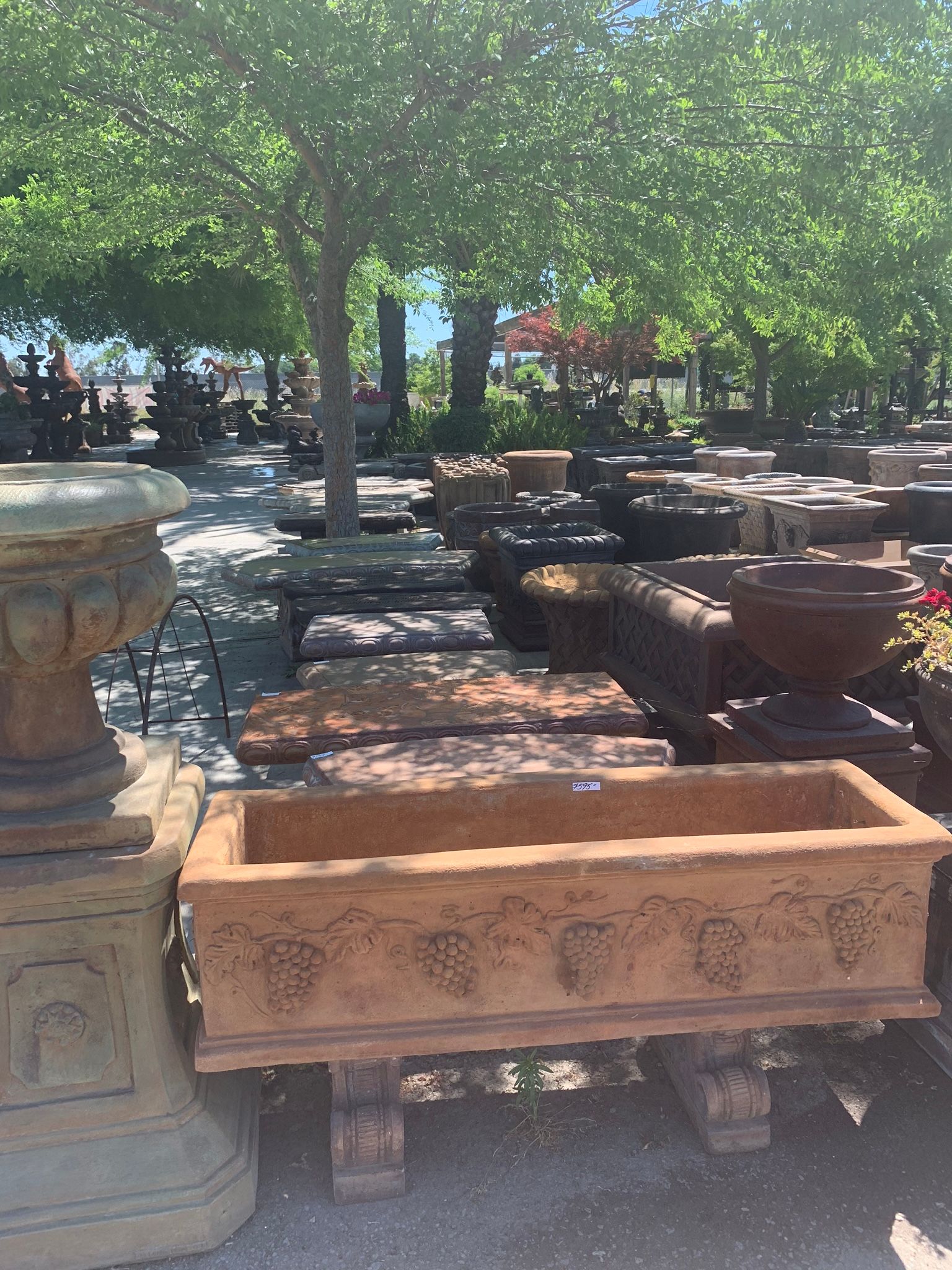 Outdoor display of stone planters and urns under trees in a garden or nursery.
