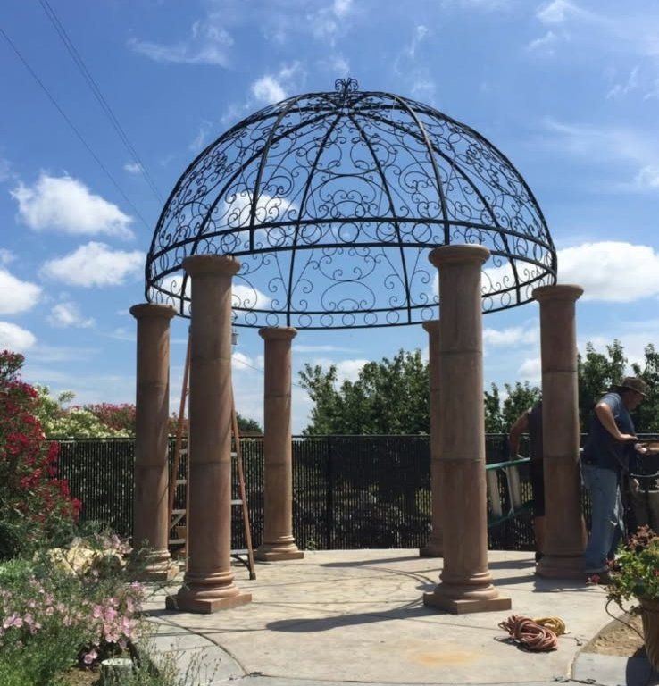 Decorative metal gazebo frame with stone columns in a sunny garden under a blue sky