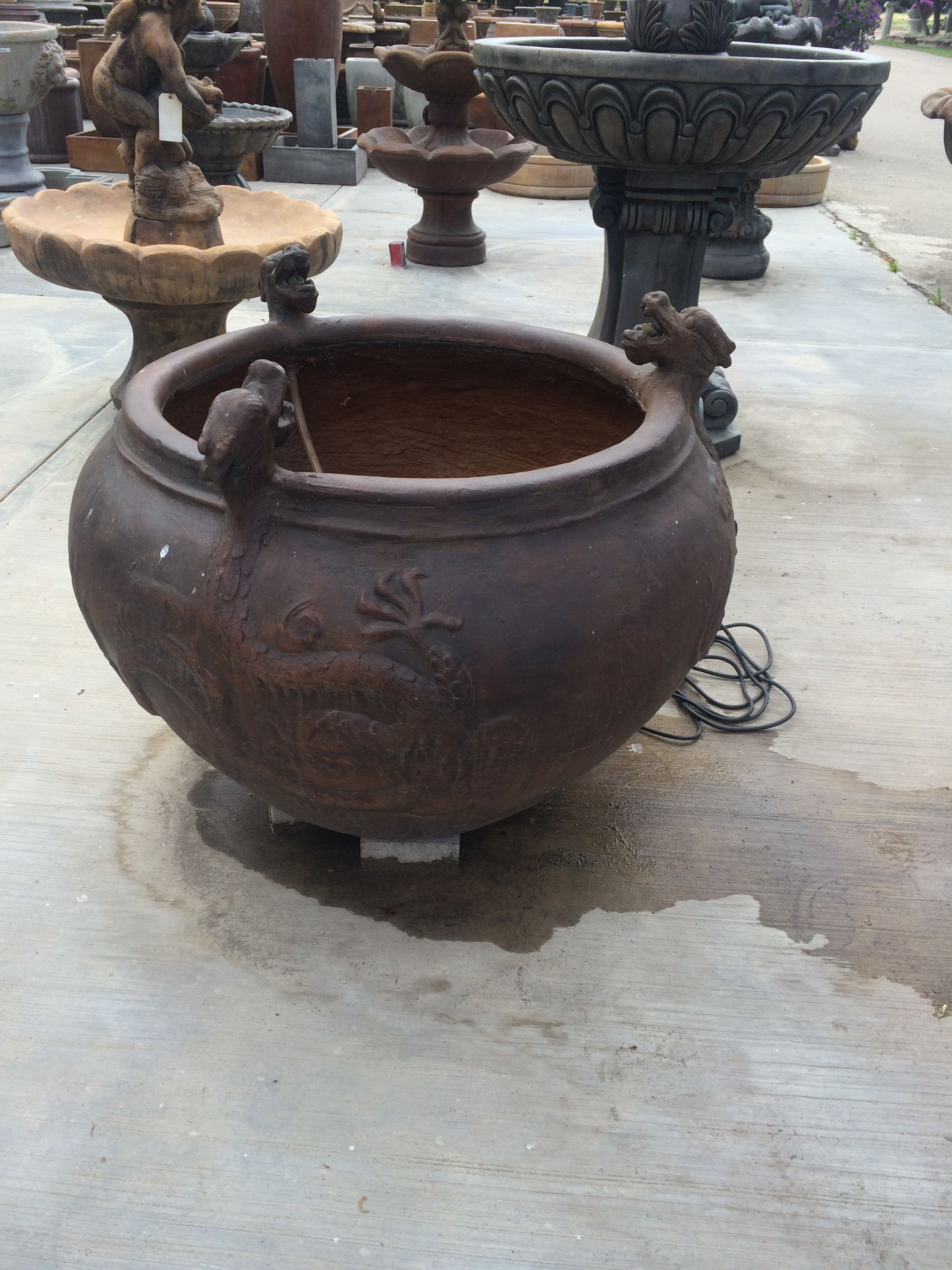 Large rust-brown metal cauldron with side handles in an outdoor courtyard, with other stone planters nearby