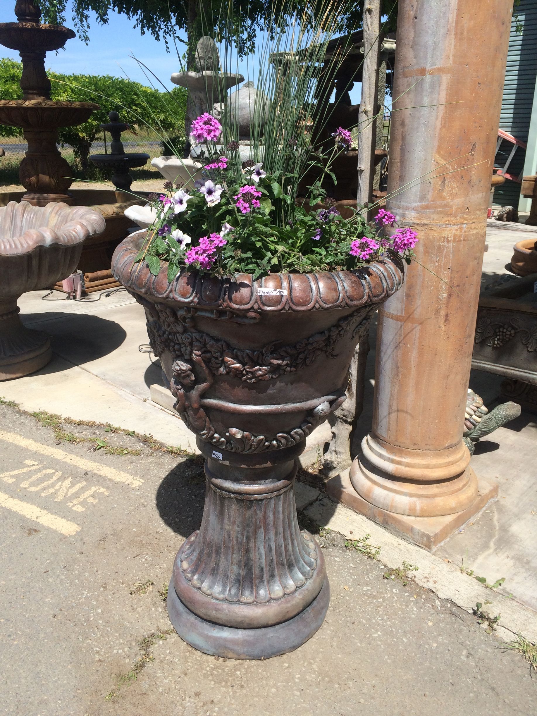 Ornate urn planter with pink flowers beside a stone column in a sunlit garden.