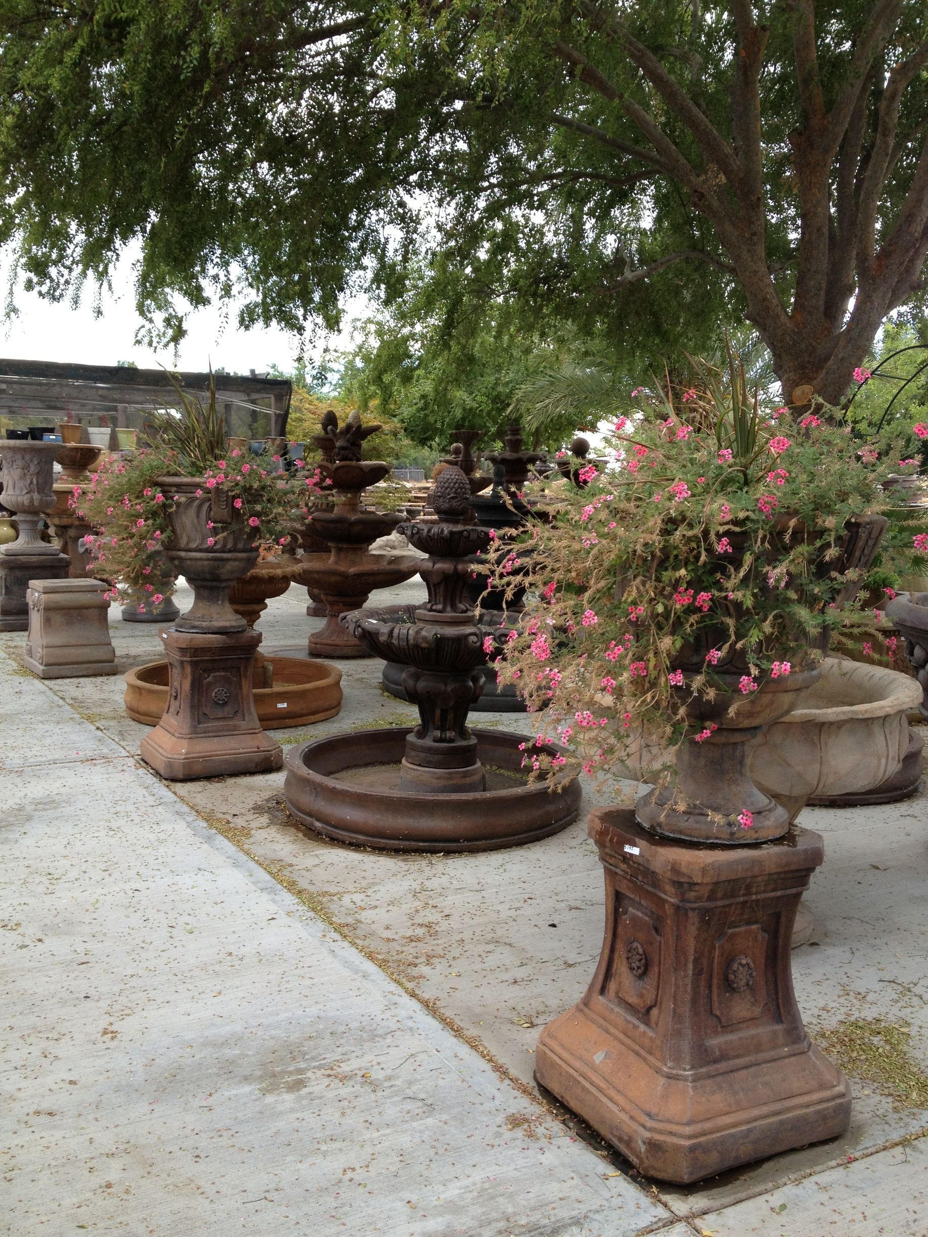 Outdoor display of stacked stone fountains and potted flowering shrubs along a garden walkway