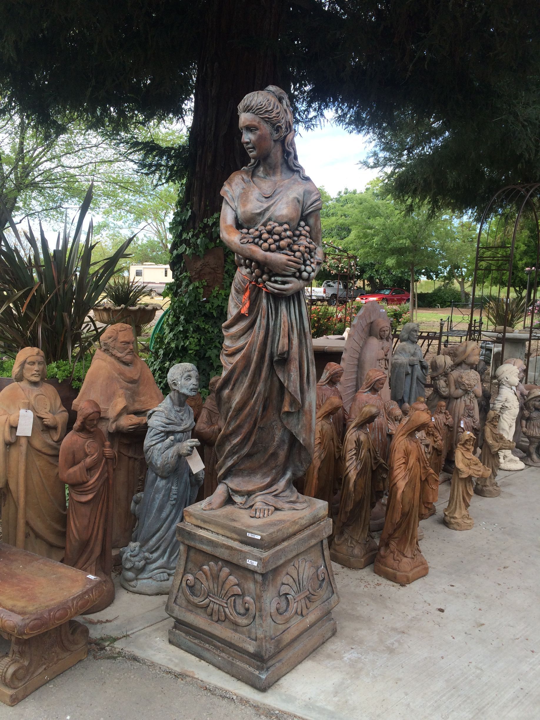 Row of weathered garden statues, centered on a tall female figure on a pedestal outdoors under trees