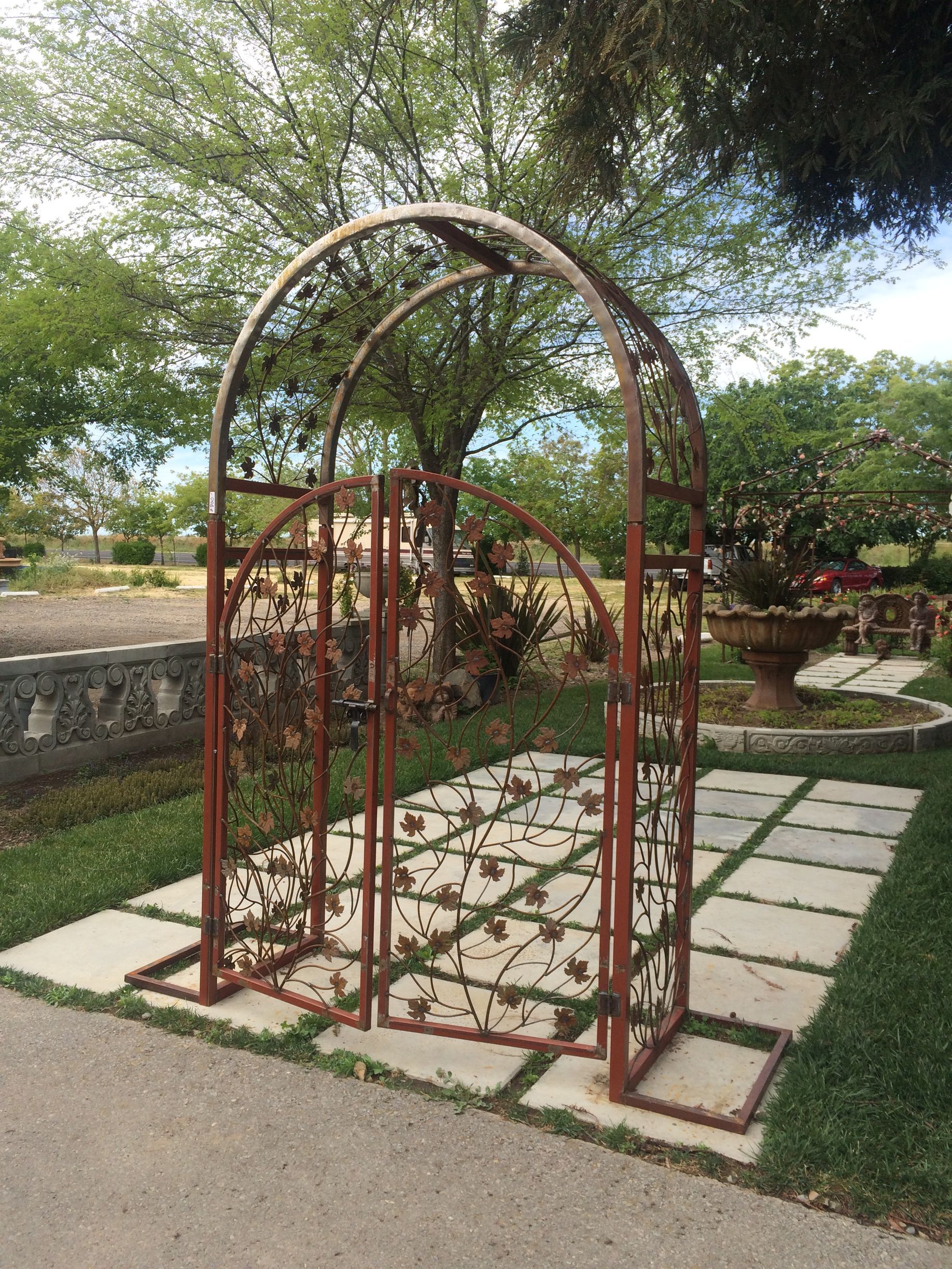 Rusty metal arch trellis over square pavers in a garden, with greenery and a stone wall behind.