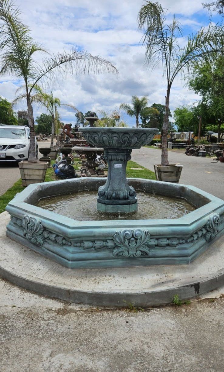 Octagonal blue fountain in a sunny plaza with palm trees and parked cars behind it