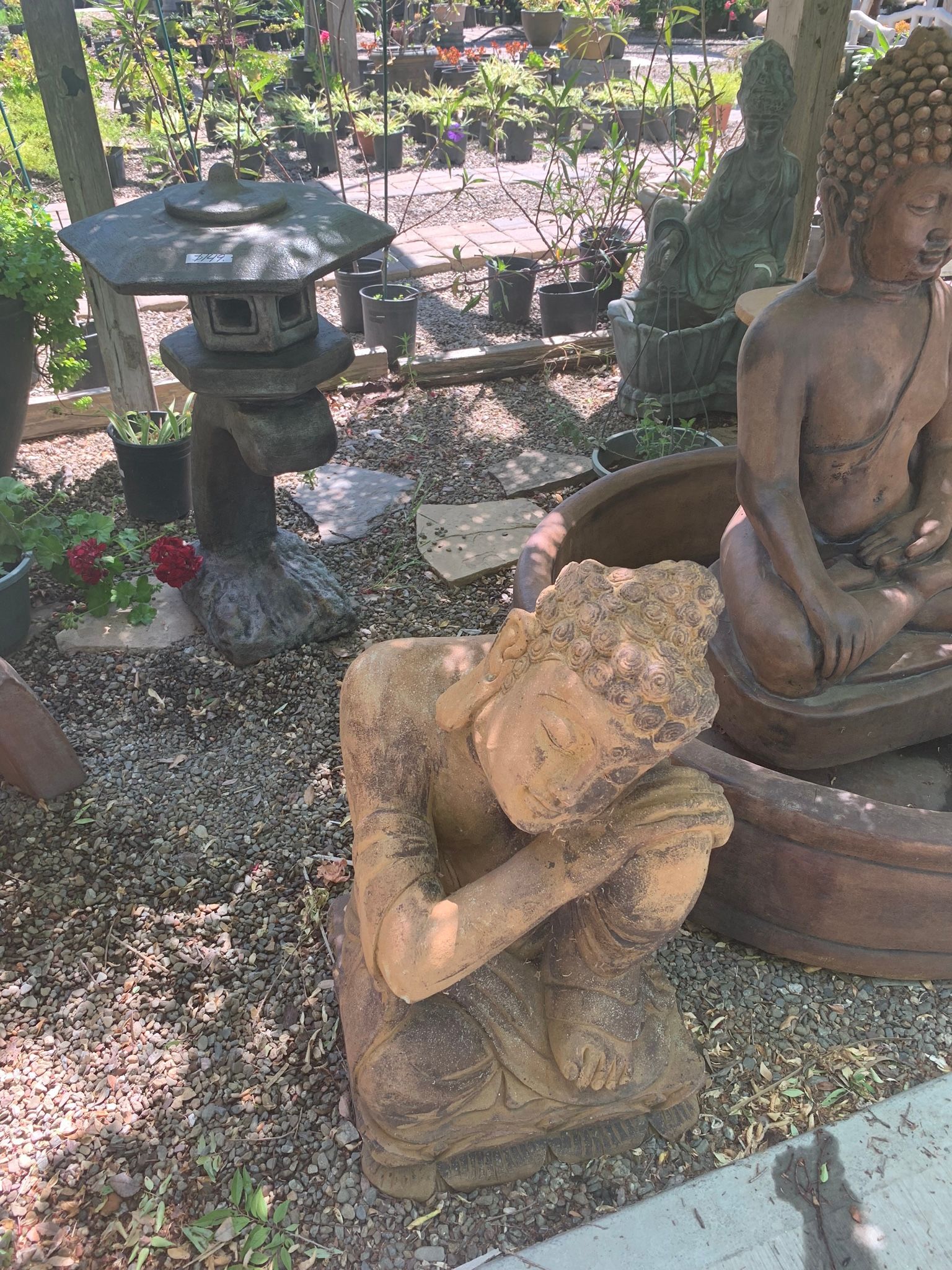 Stone garden statues among plants and gravel, including a kneeling figure in the foreground and seated statue at right