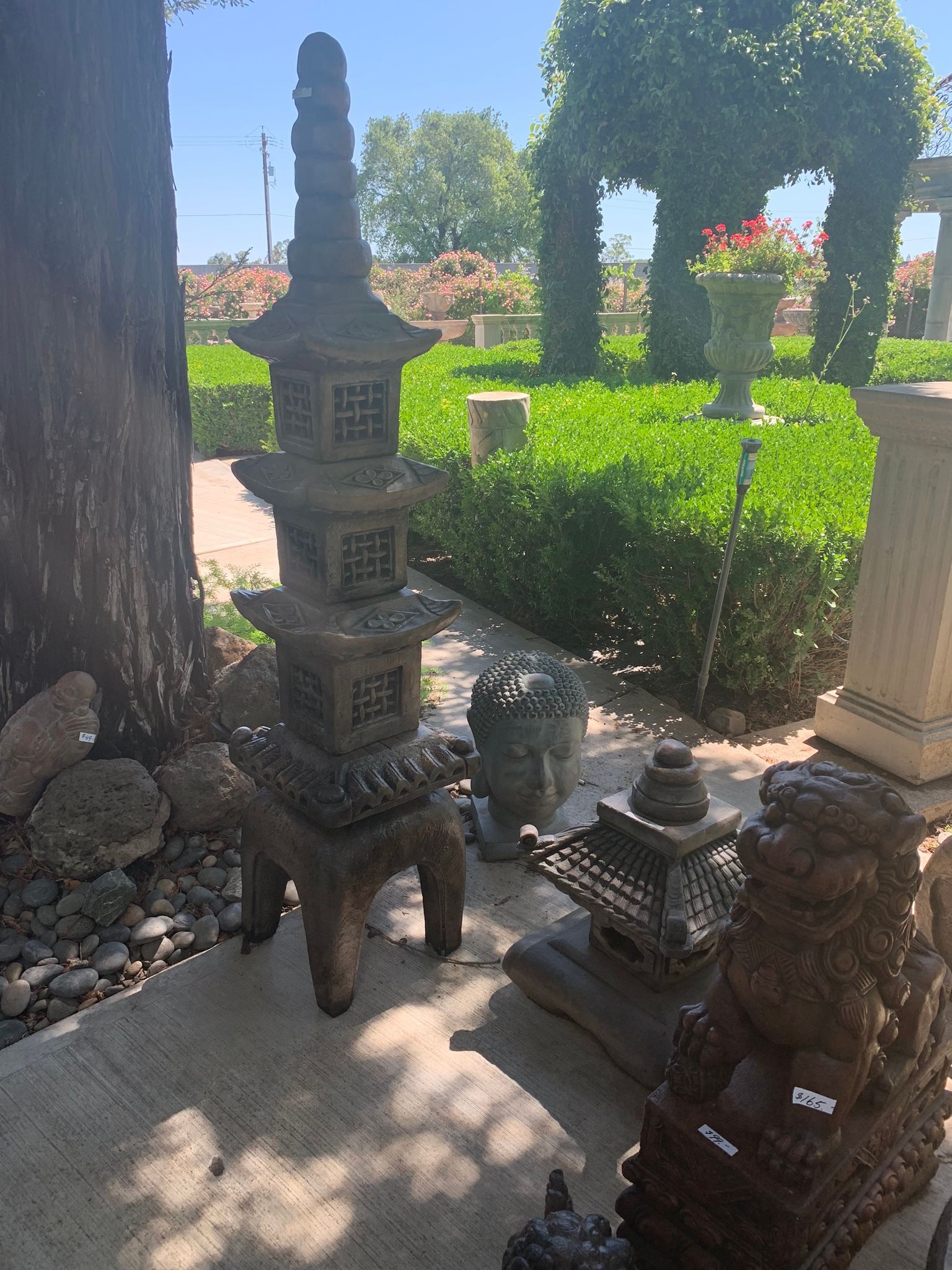 Stone garden statues and carved lanterns beside a hedge in a sunny outdoor courtyard