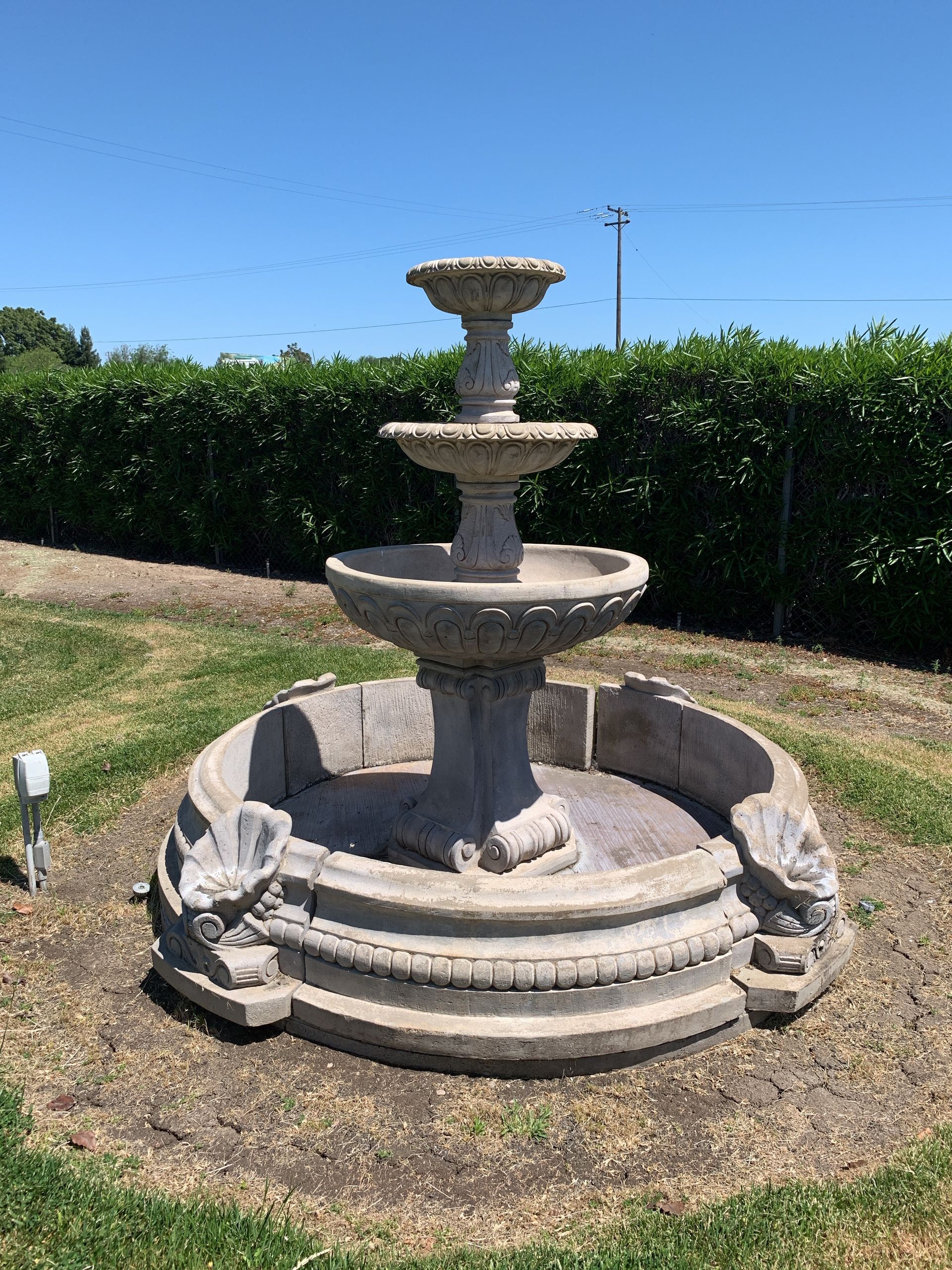 Stone tiered fountain in a grassy garden, with hedges and blue sky in the background.