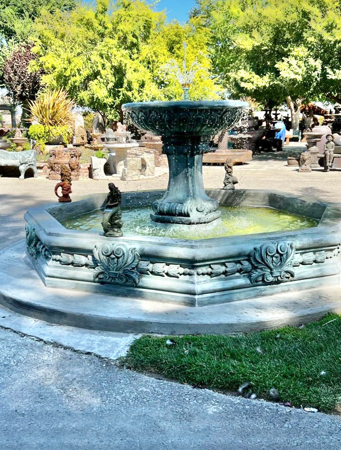 Stone fountain in a sunny park, surrounded by trees and benches.