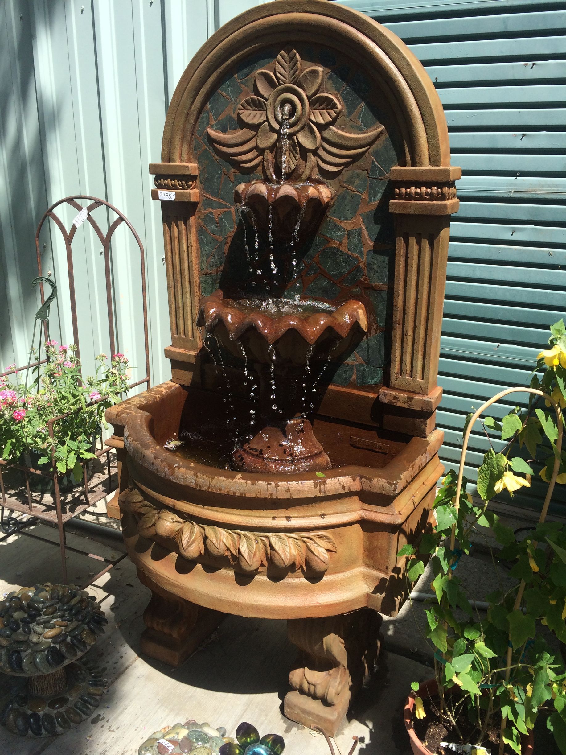 Ornate terracotta fountain with running water beside corrugated metal walls and potted plants
