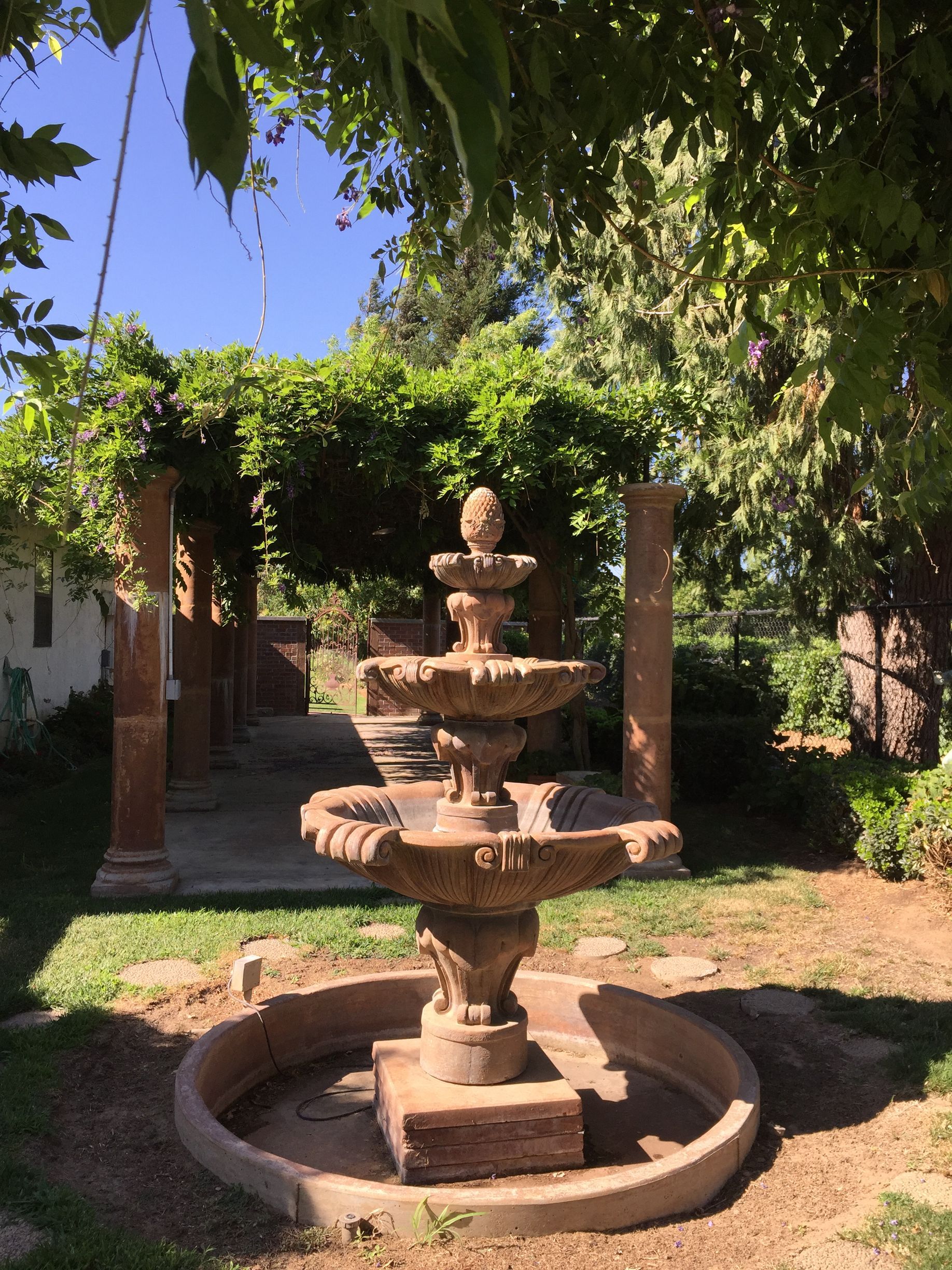 Tiered stone fountain in a shaded garden courtyard with brick columns and greenery