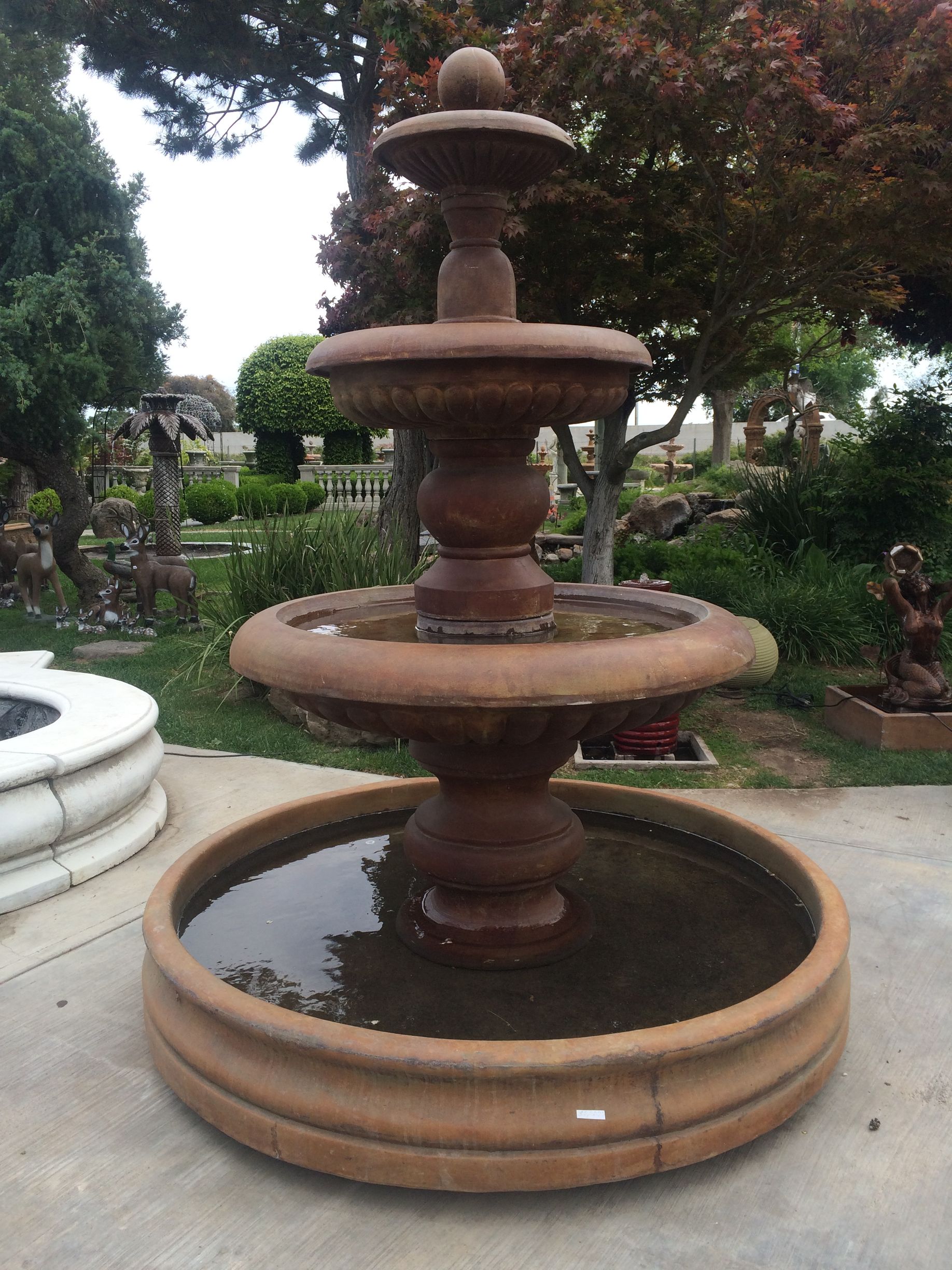 Tiered stone fountain in a garden, with reddish-brown basins and greenery in the background