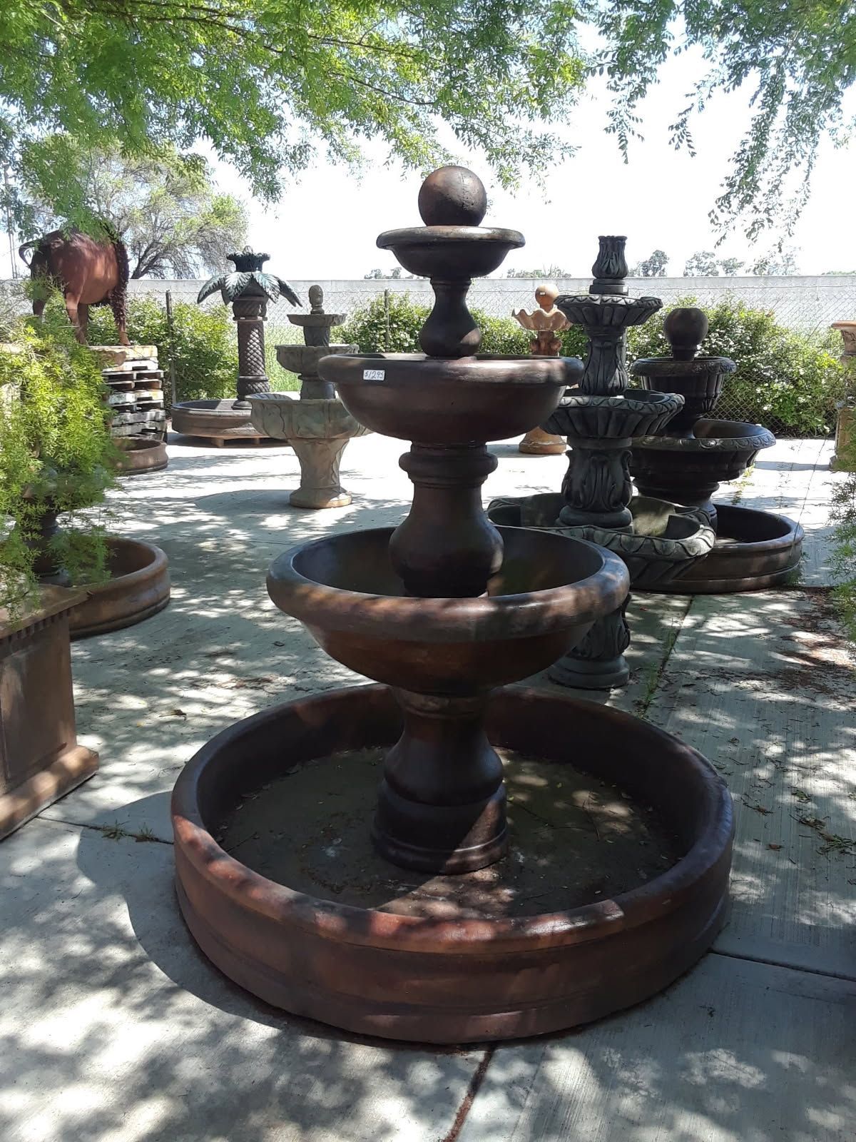 Tiered stone fountain in a sunlit garden with trees and potted plants around it