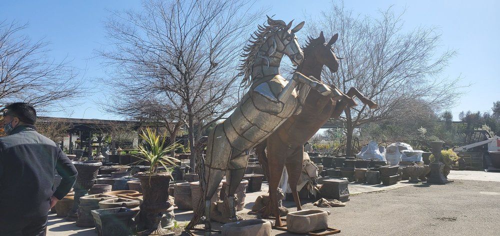 Large horse statue in a sunny outdoor garden, with potted plants and a person standing at left