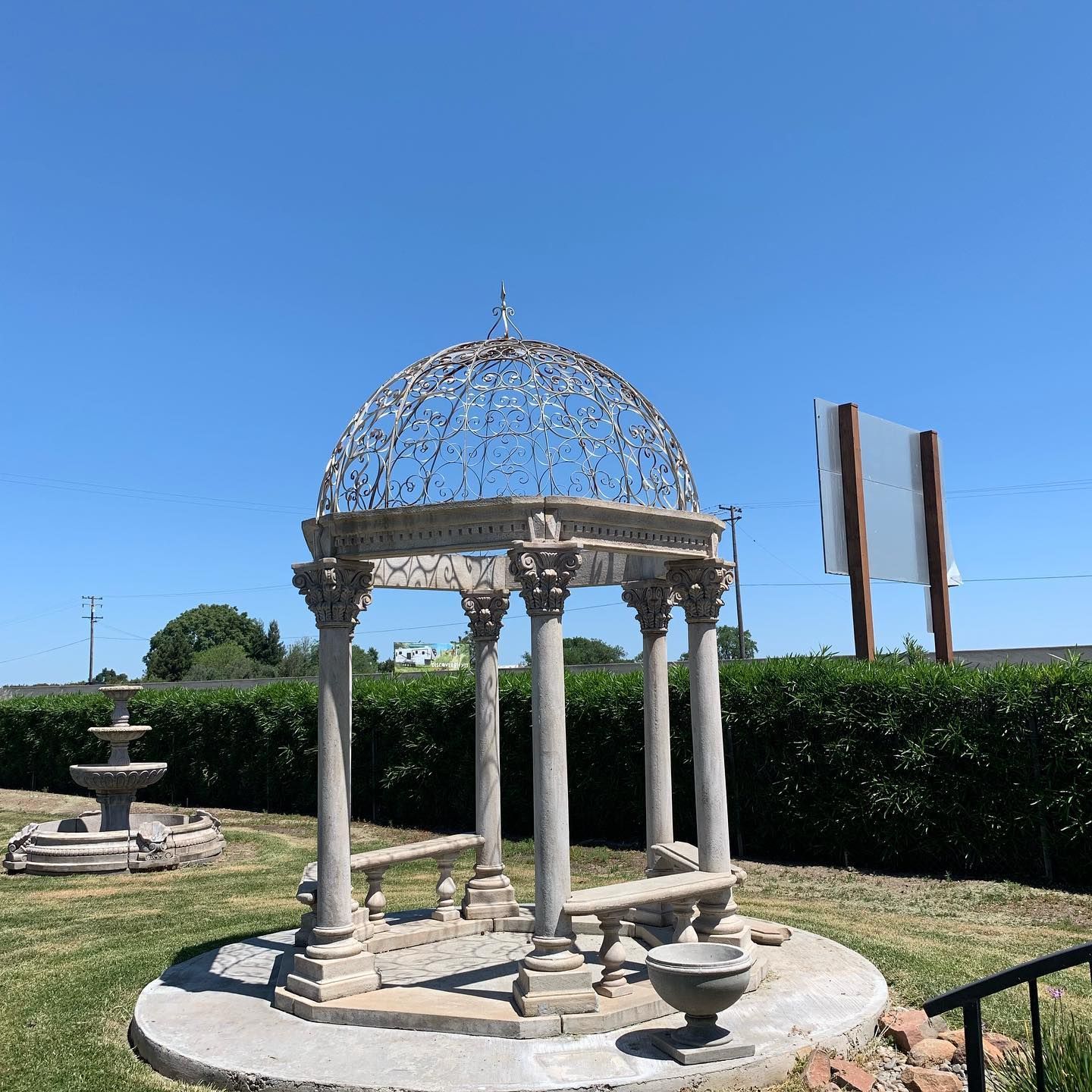 Small stone gazebo with columns and a domed metal roof in a sunny garden, with hedges and fountain basins nearby