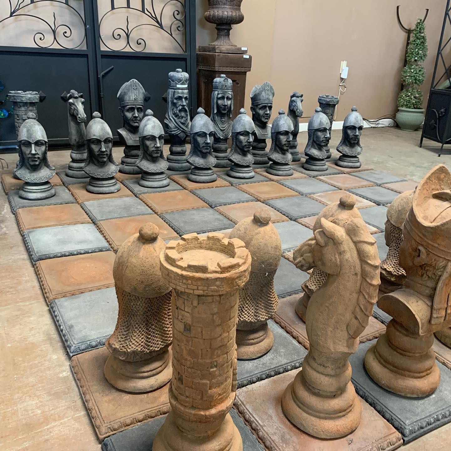 Outdoor chessboard with oversized stone chess pieces arranged on a tiled patio near a gate.