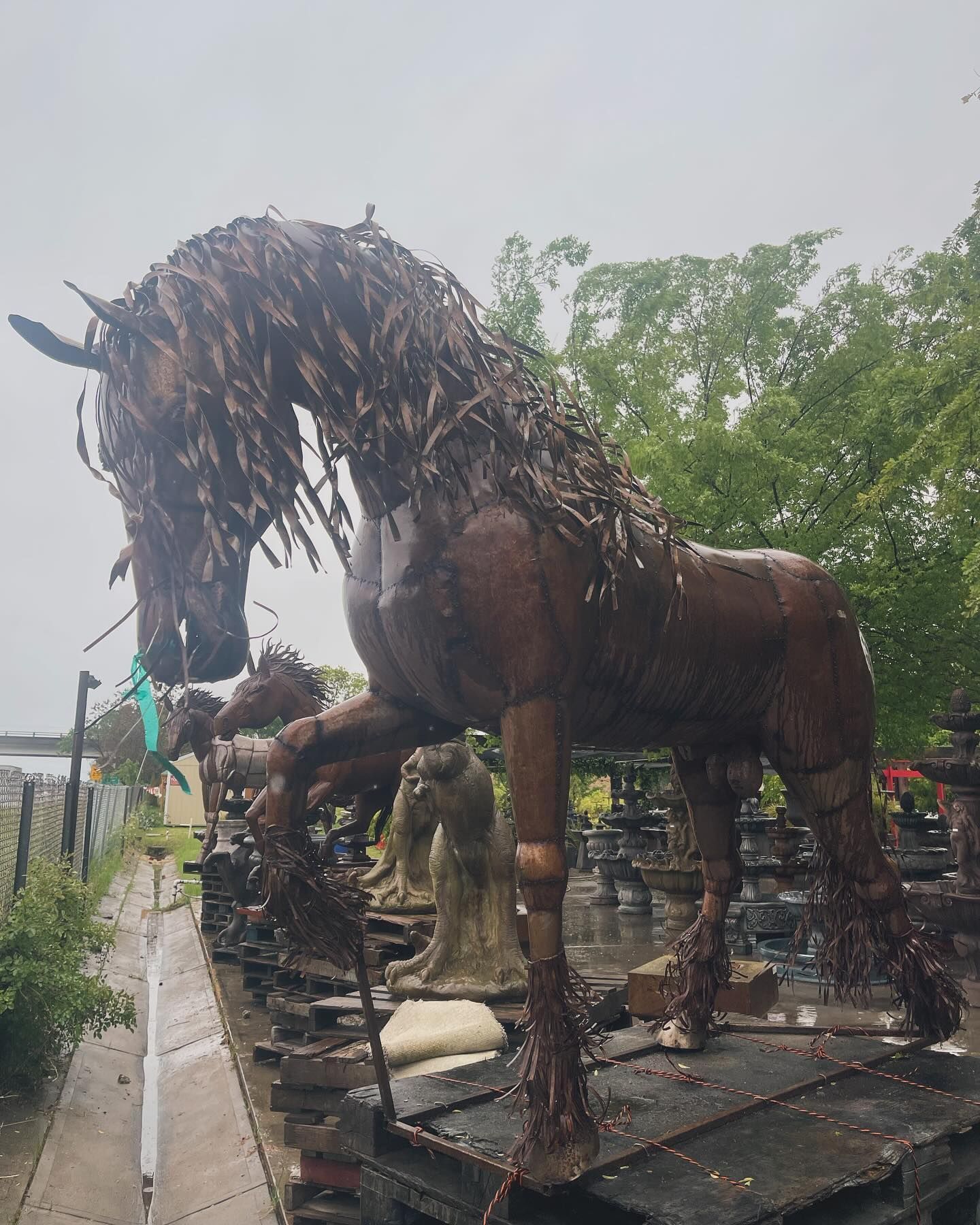 Large rusted horse sculpture on a platform outdoors, with trees and a cloudy sky in the background.