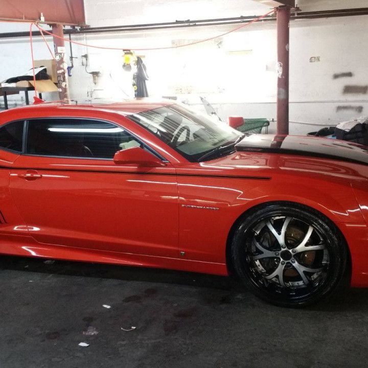 Red Chevrolet Camaro with black hood and racing stripe parked in a garage.