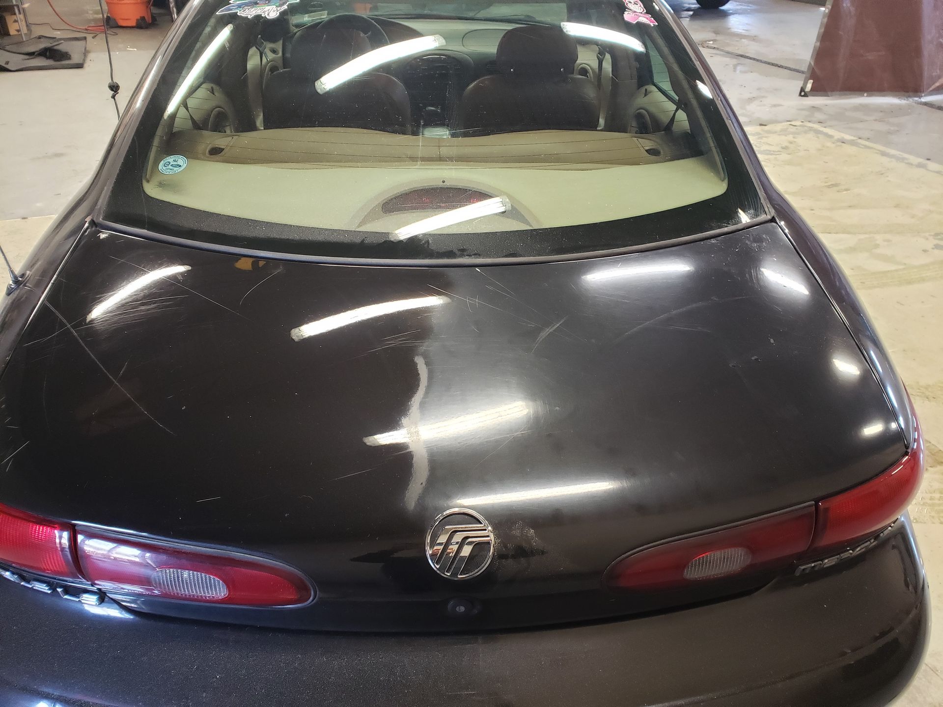 Dark car's rear, scratched paint, red taillights, round emblem, in a garage.