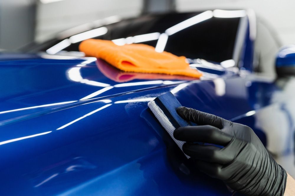 A gloved hand uses a foam block to apply ceramic coating to the blue hood of a car with an orange towel nearby.