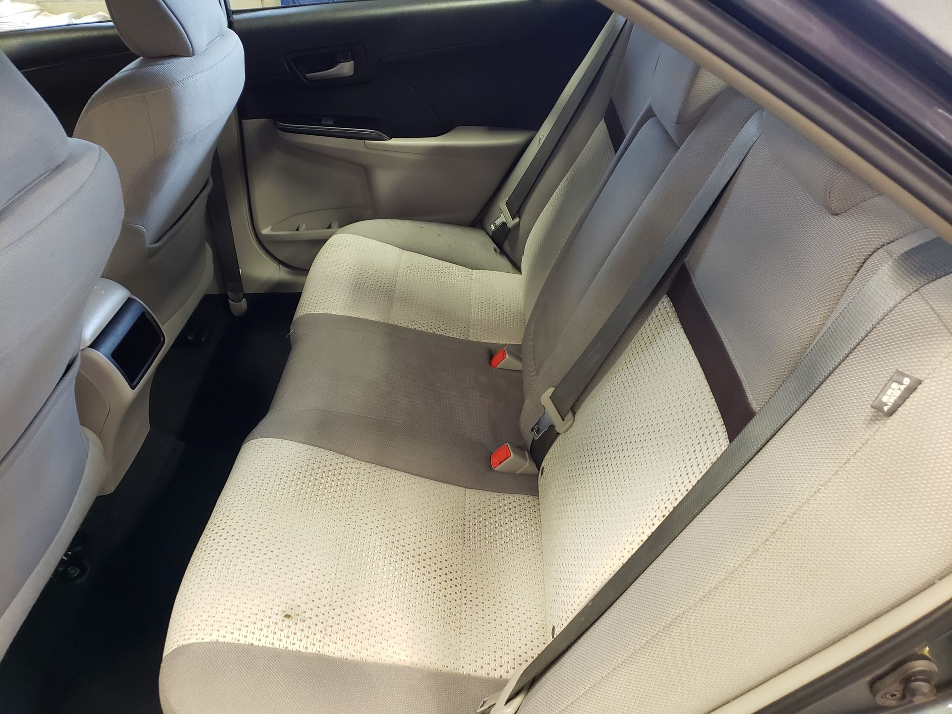Back seat of a car, with gray and white cloth upholstery and seat belts.