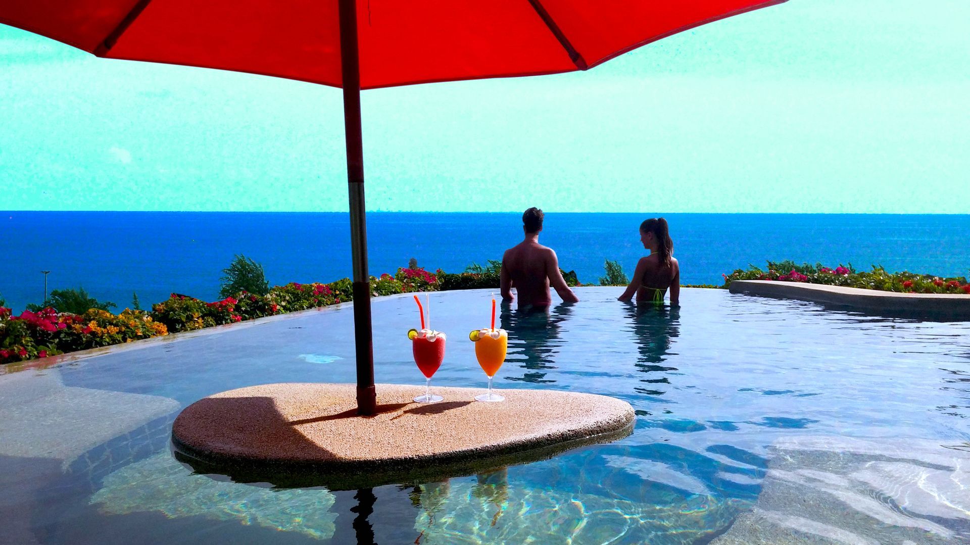 A couple sitting in a swimming pool with drinks under an umbrella
