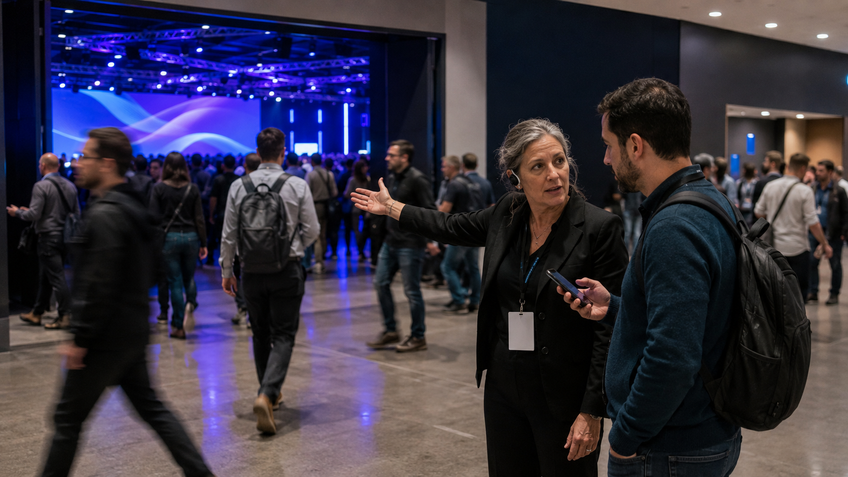 Crowd in a lobby with blue-lit stage screens; two people talk in the foreground.