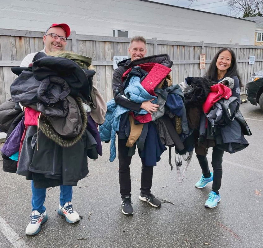 Three people are holding a pile of clothes in a parking lot