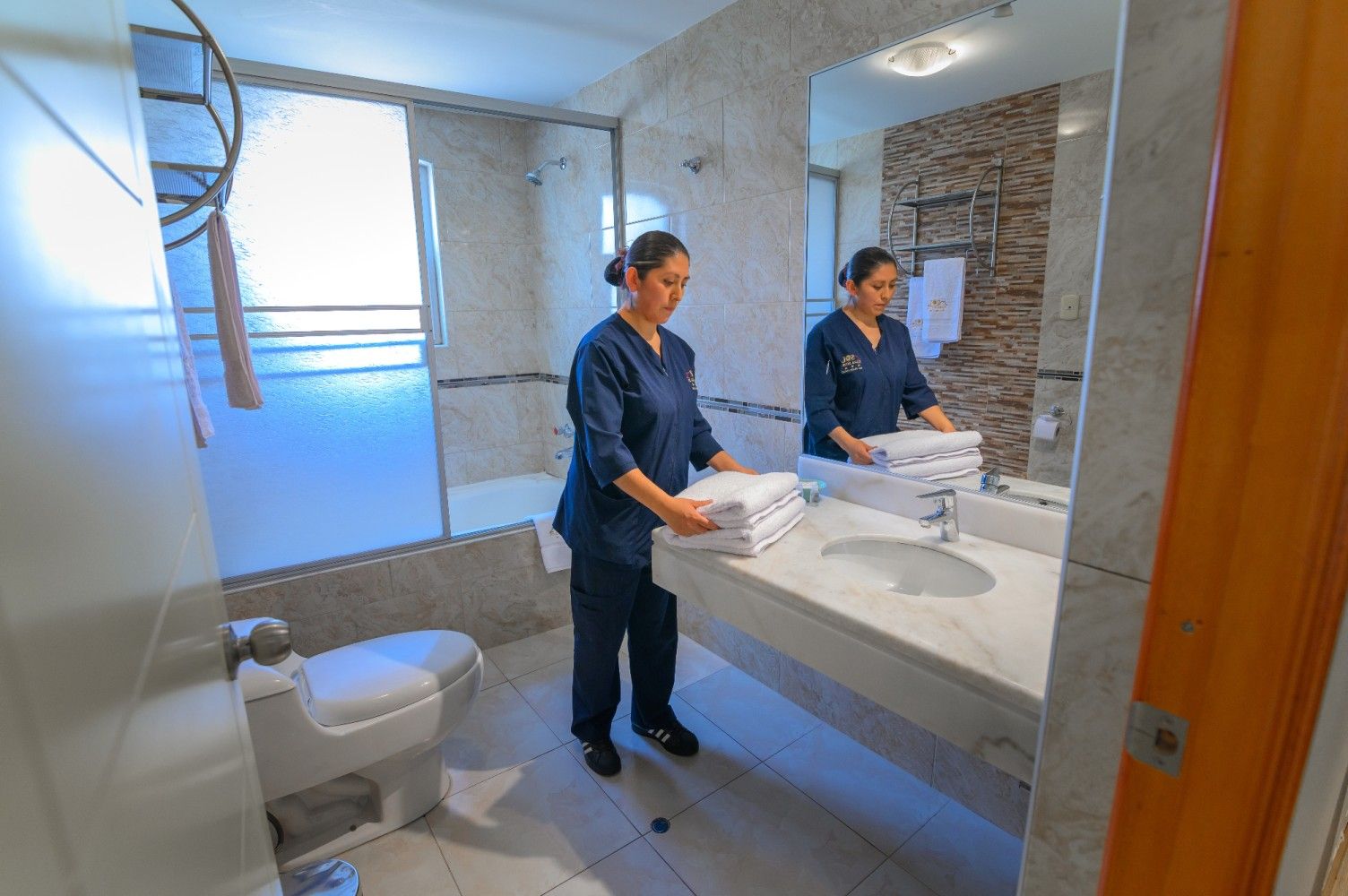 Un miembro del personal, vestido con uniforme azul, coloca toallas blancas dobladas sobre el mostrador de mármol del baño.