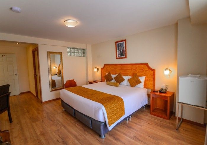Una habitación de hotel con cama de matrimonio, alfombra dorada, suelo de madera, lámparas de noche y un pequeño frigorífico.