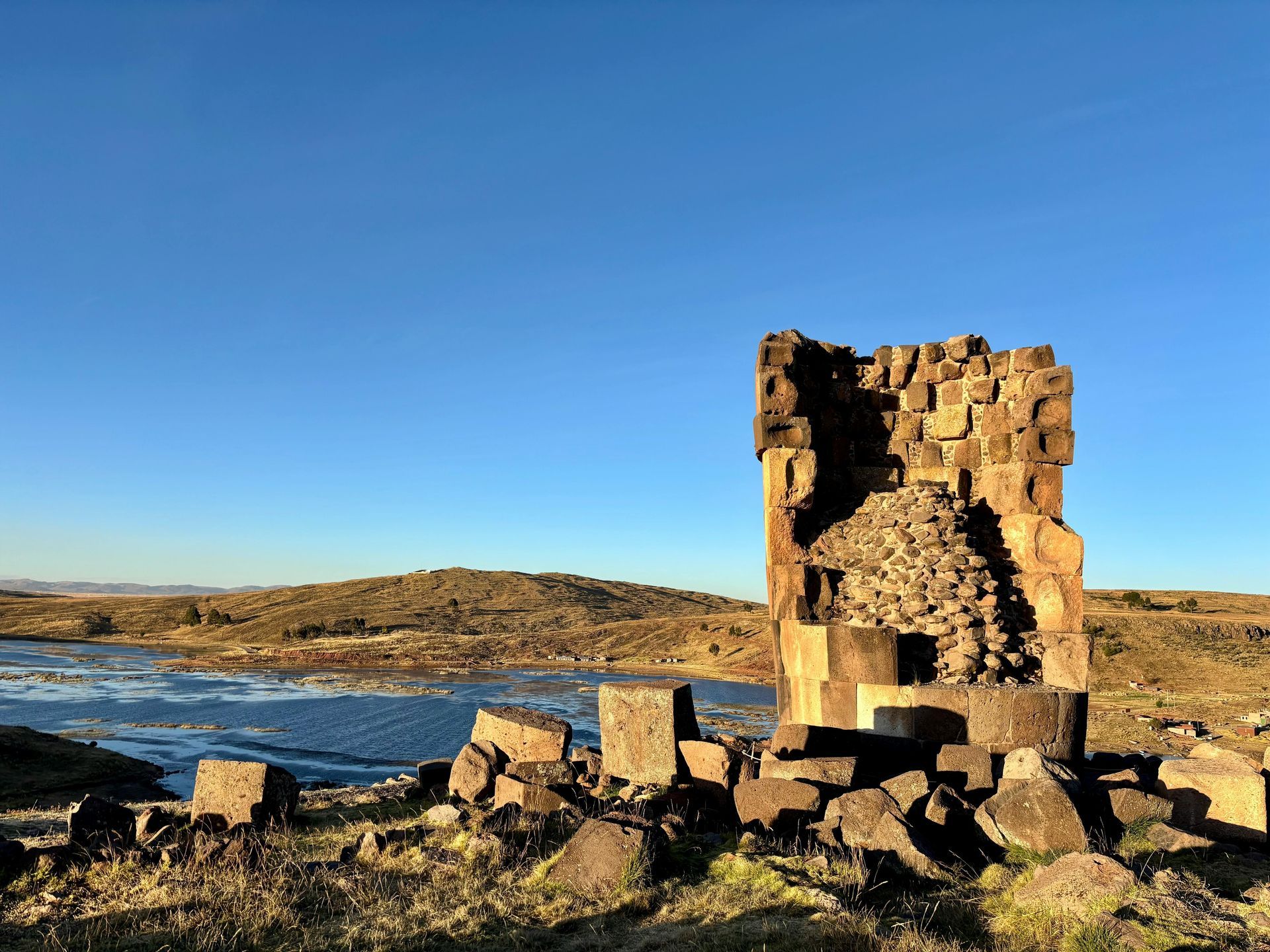 Antiguas ruinas de piedra se alzan cerca de un lago azul bajo un cielo claro y brillante, en un paisaje rocoso y árido.
