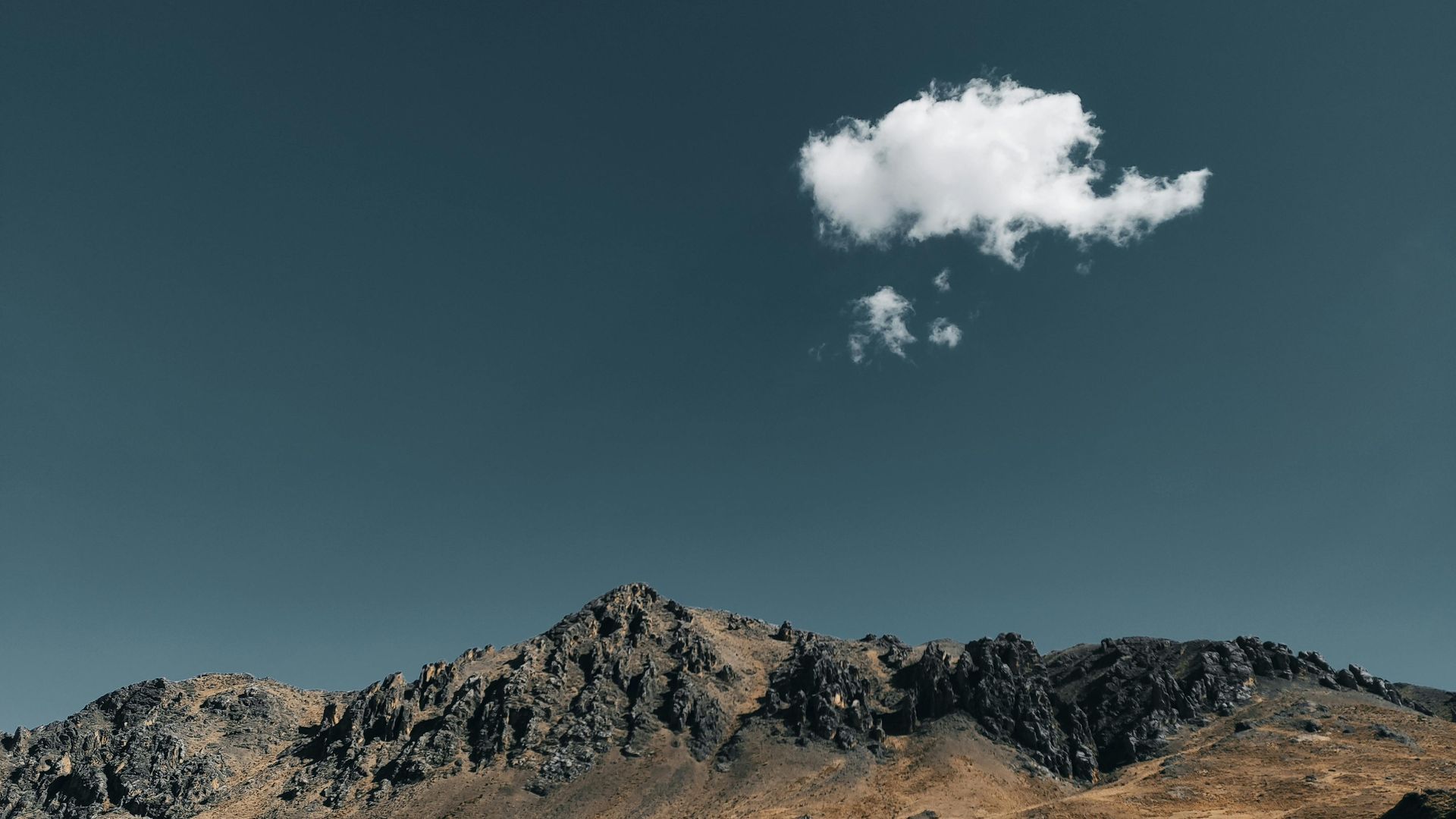 Una solitaria nube blanca flota sobre una oscura y escarpada cordillera, recortada contra un cielo azul intenso.
