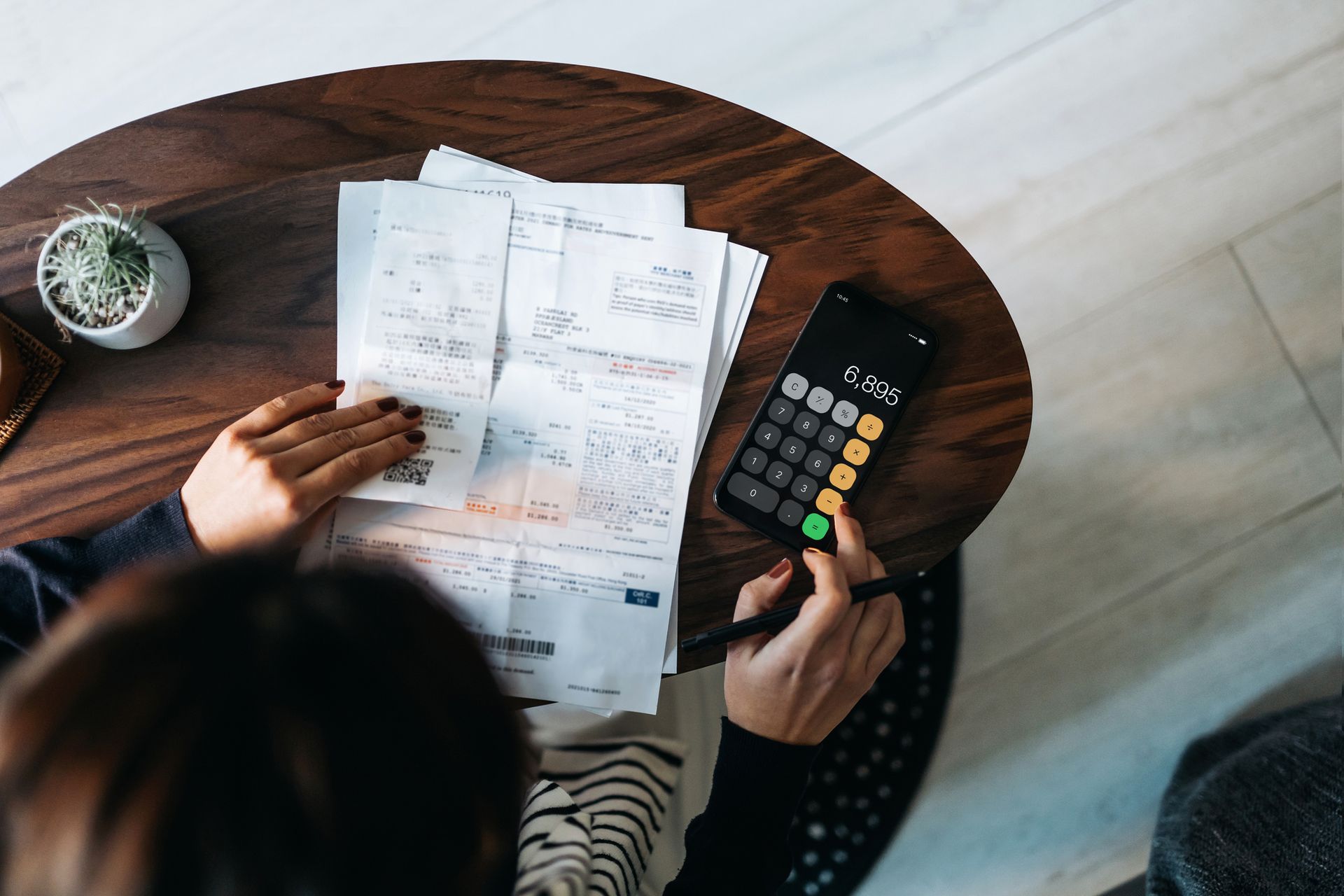 Person calculating expenses on phone, surrounded by bills on a wooden table.