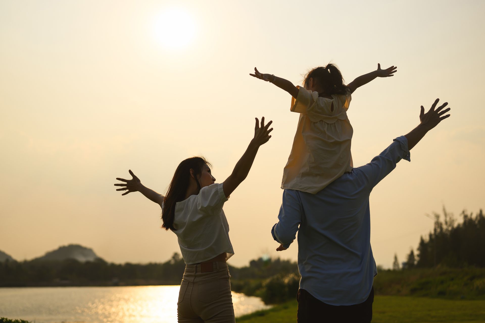 Family with arms raised at sunset by a lake; a child sits on the father's shoulders.