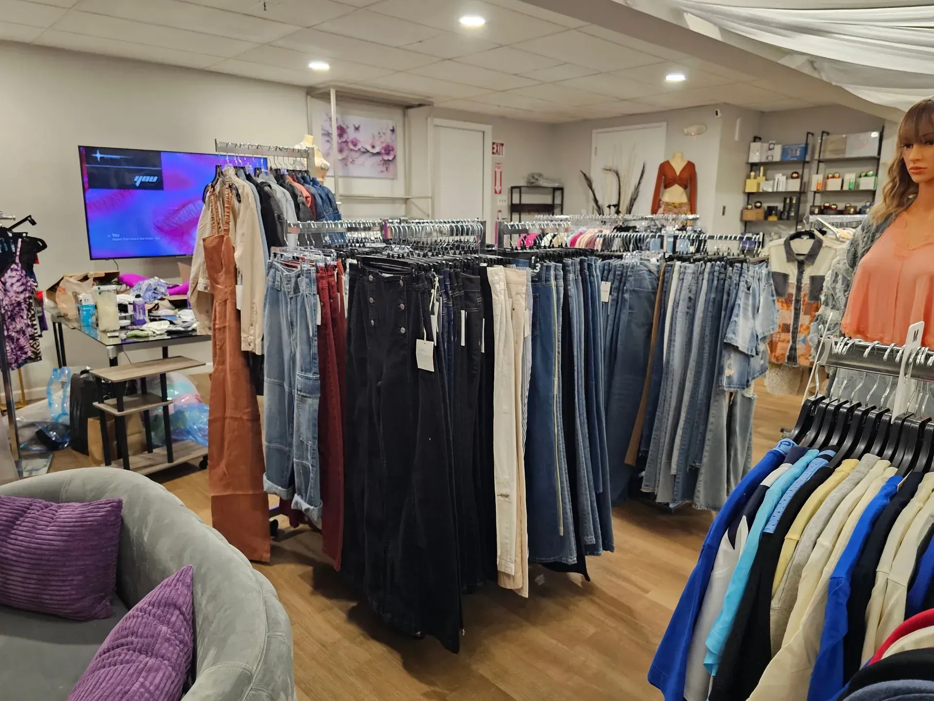 Clothing boutique with racks of jeans and dresses, a TV, and a gray chair in a bright room