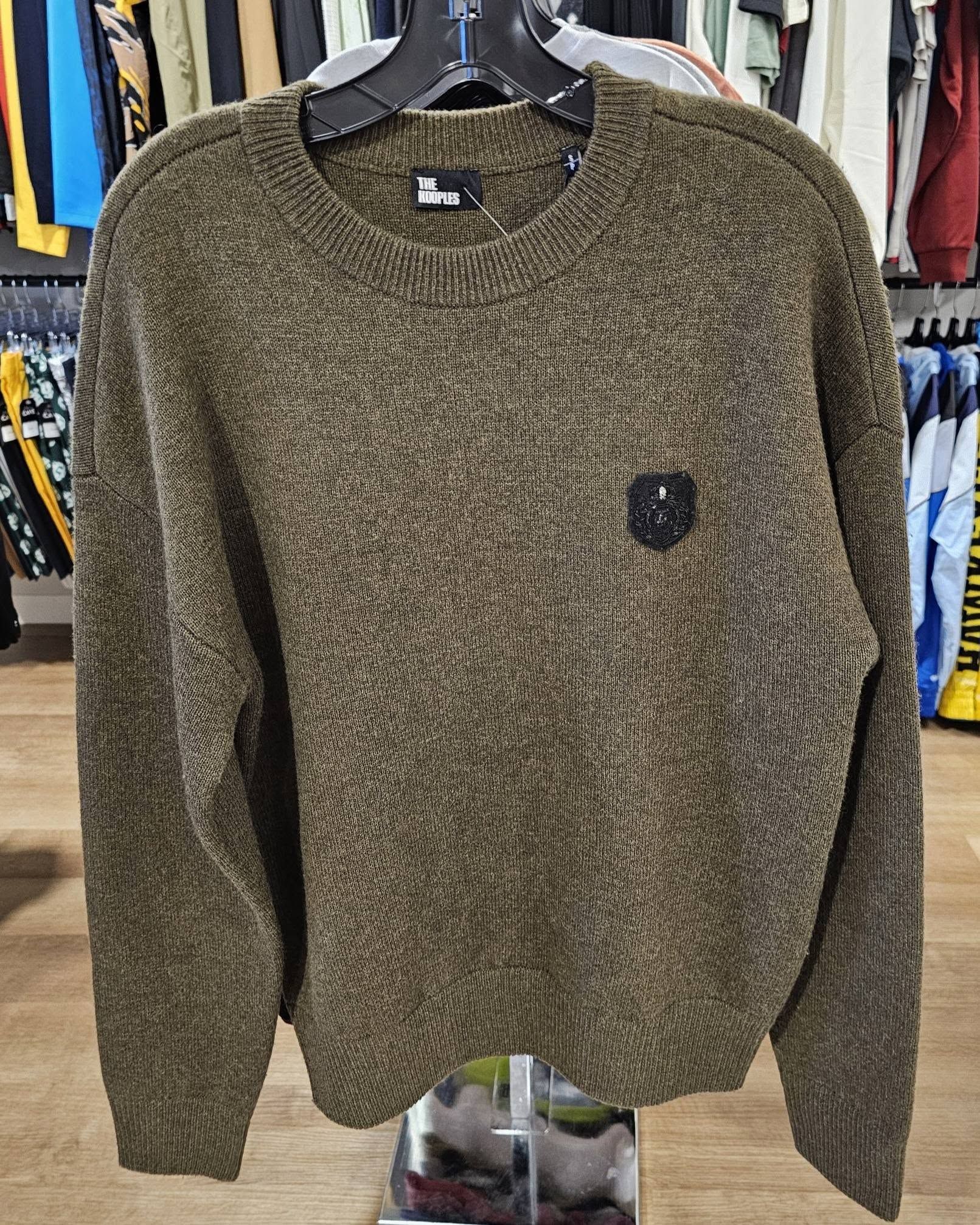 Olive green knit sweater with a small chest patch, hanging on a rack in a clothing store.