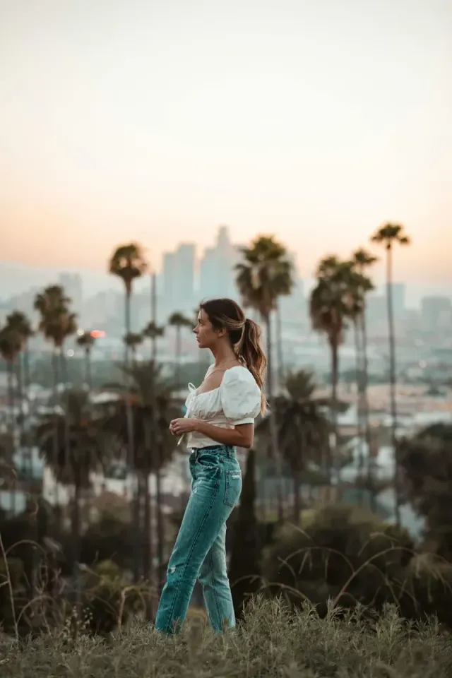 Woman standing in grass at sunset with palm trees and city skyline in the background