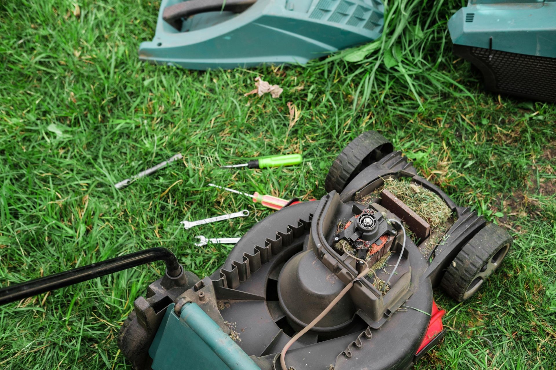 Damaged green lawn mower on grass with scattered tools and exposed engine parts